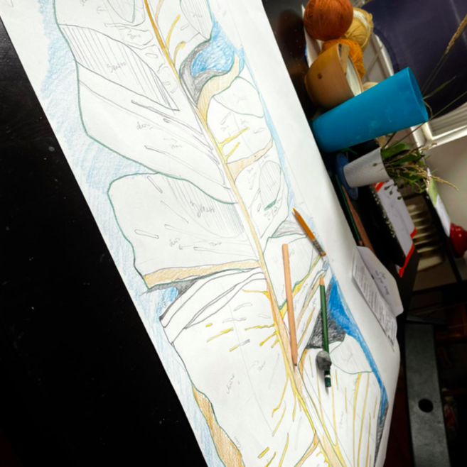 Colorful botanical illustration of a leaf with detailed vein structure on a workspace table surrounded by art supplies.