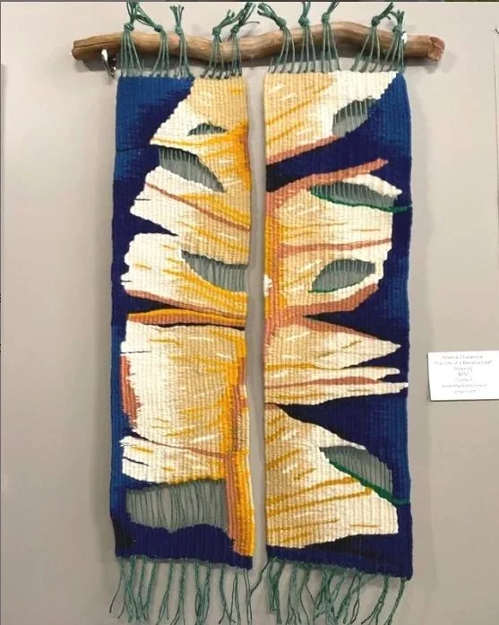 Colorful textile wall hanging resembling a sliced banana, with green leaves on a dark blue background, attached to a wooden rod.