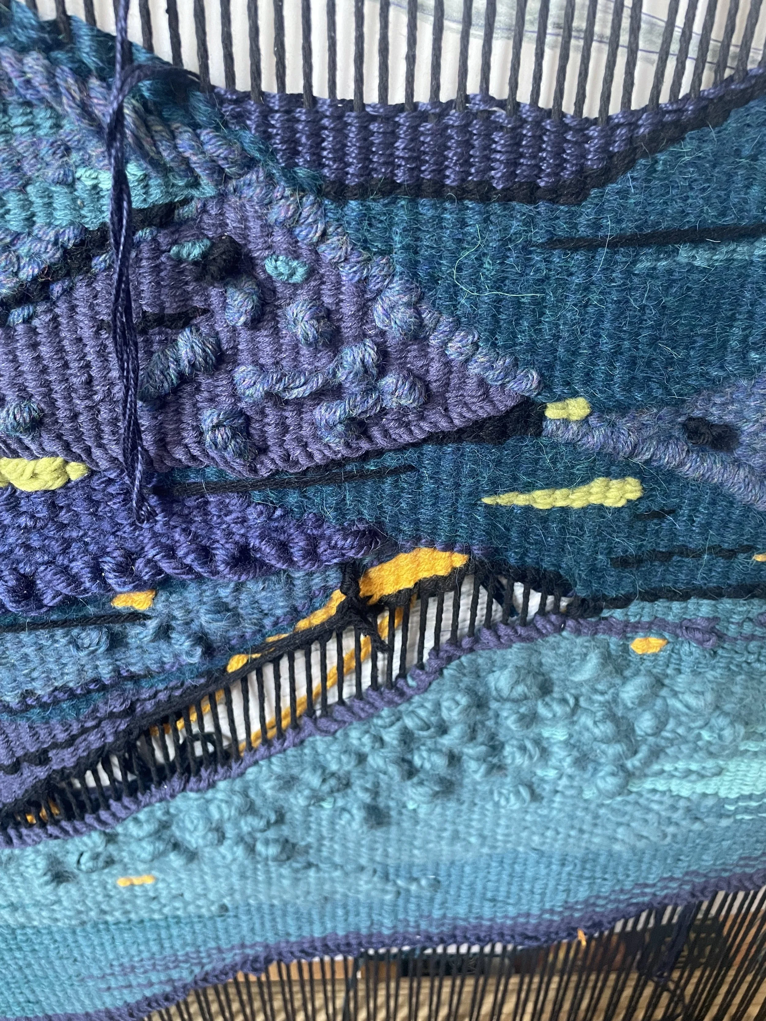 Close-up of colorful, textured woven fabric with purple, teal, and yellow yarns on a black wire loom.