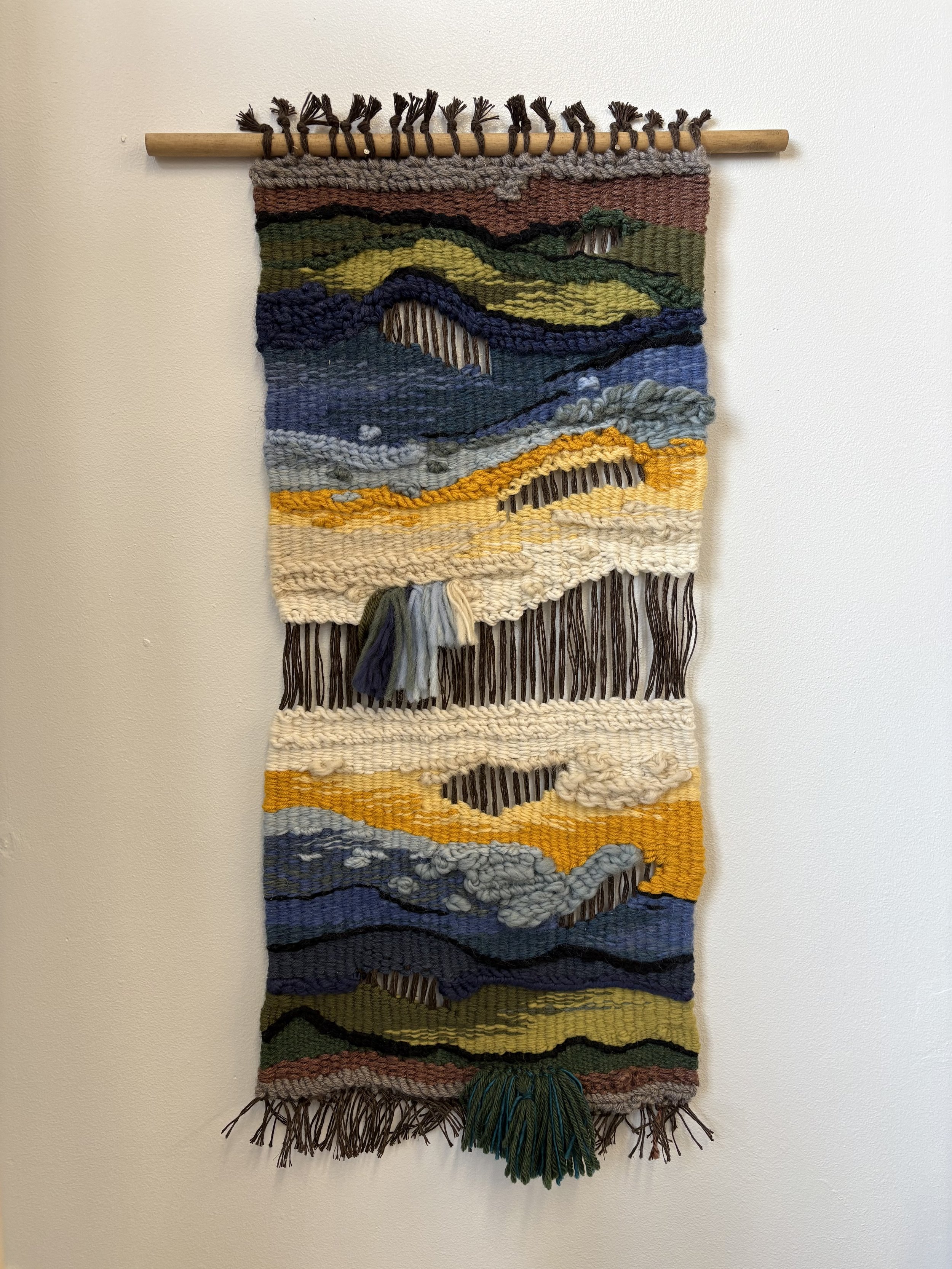 Colorful woven tapestry depicting mountains, sky, and landscape, hanging on a wall.