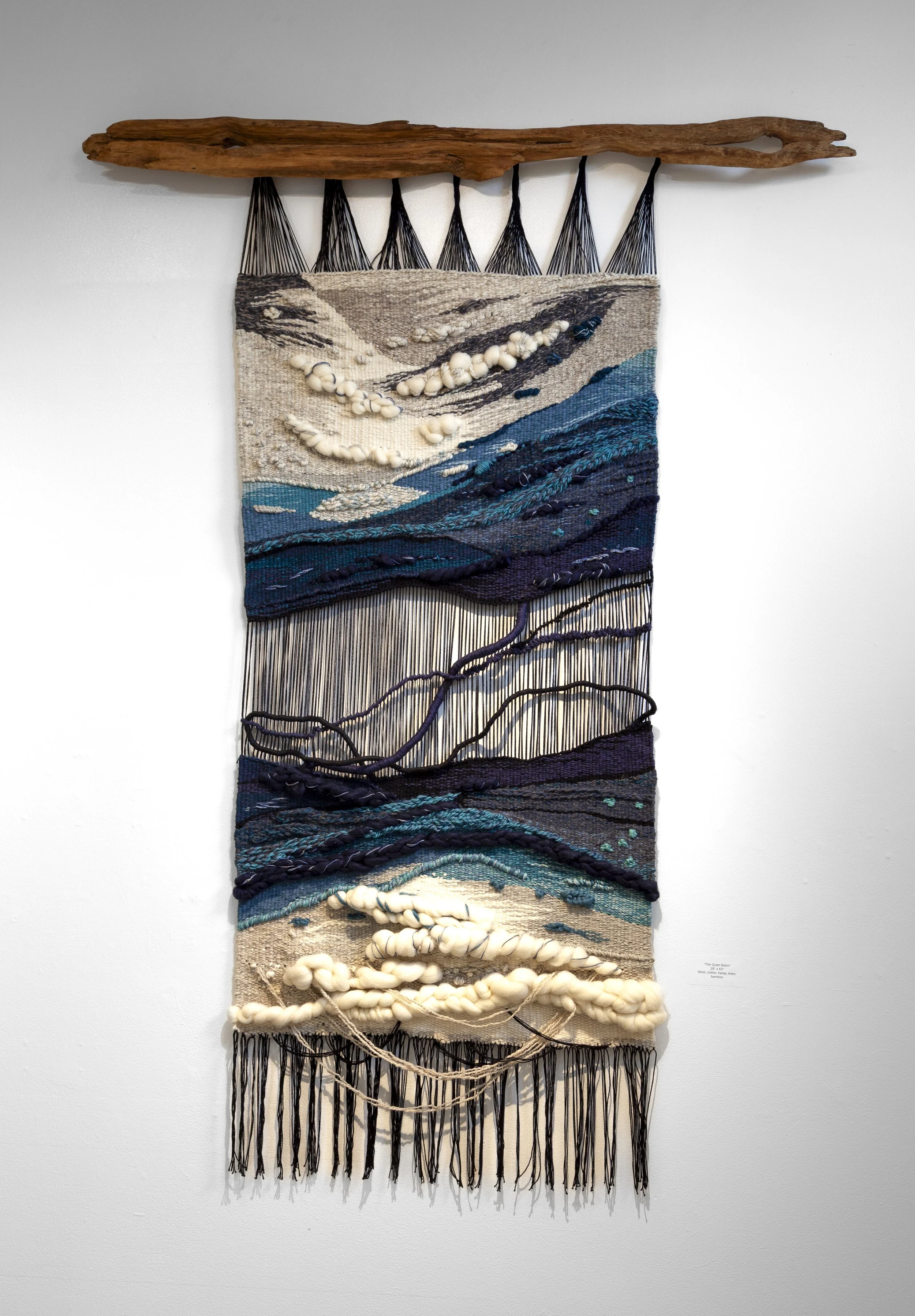 A textile wall hanging with abstract wave patterns in shades of blue, black, cream, and beige, featuring textured loops and fringe at the top and bottom, mounted on a white wall.