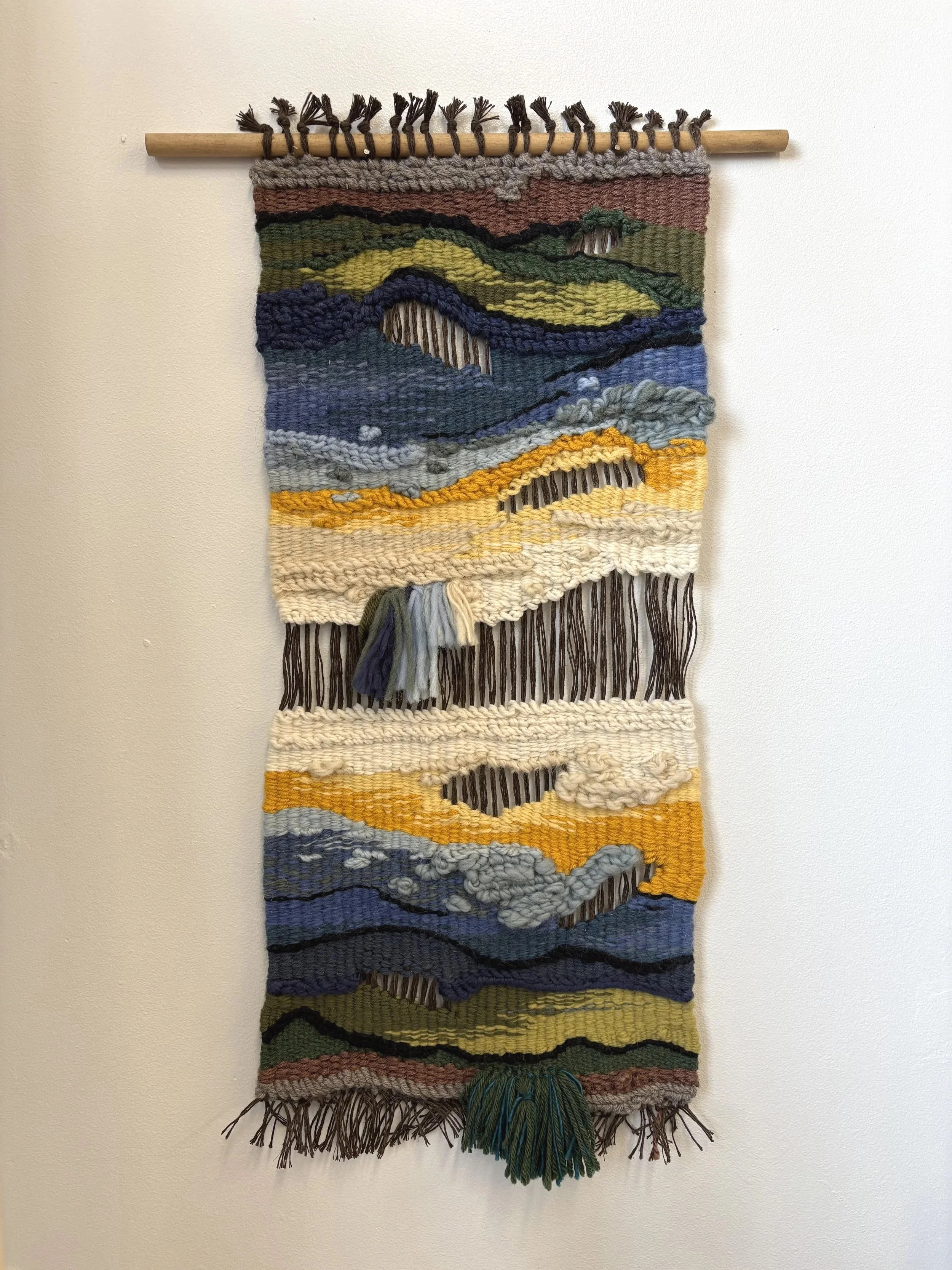 Colorful woven wall hanging depicting a landscape scene with mountains, trees, and a sunset.