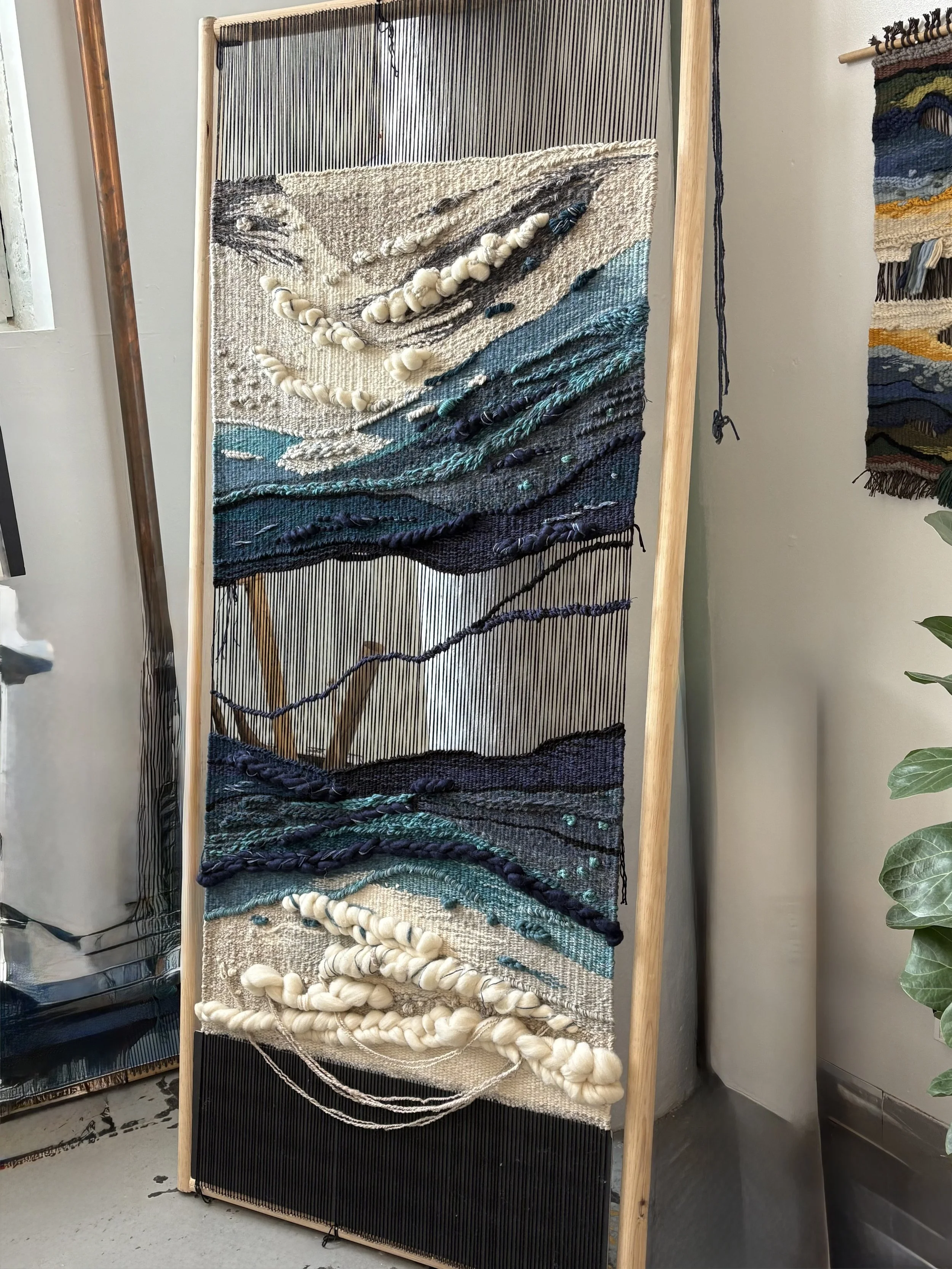A textile art piece on a wooden frame with abstract waves, made from woven and chunky yarns in shades of white, cream, blue, and black.