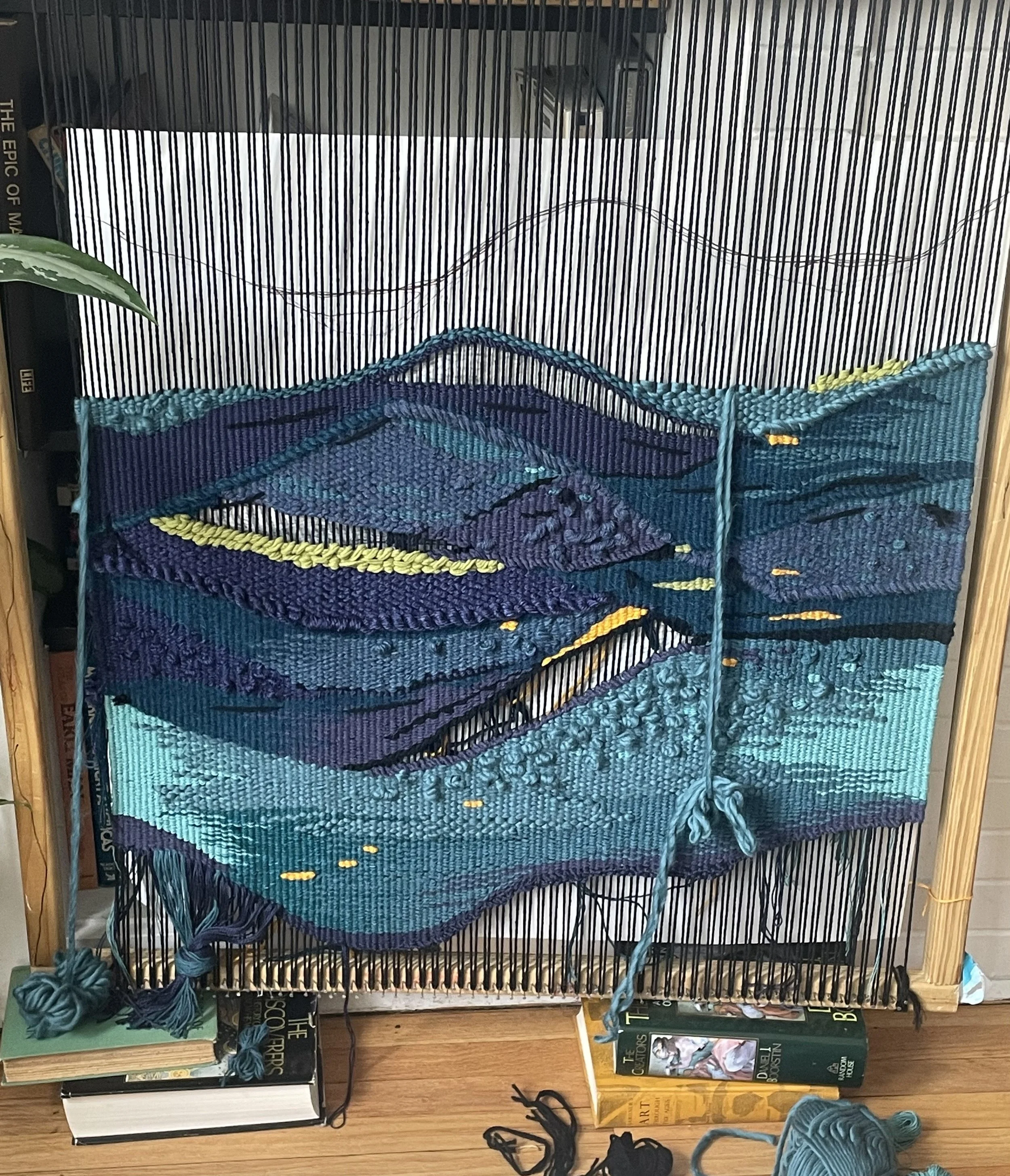 A framed textile art piece made from woven yarns in shades of blue and purple, depicting a landscape scene with mountains, hills, a river, and a yellow pathway, displayed on a wooden table with books underneath.