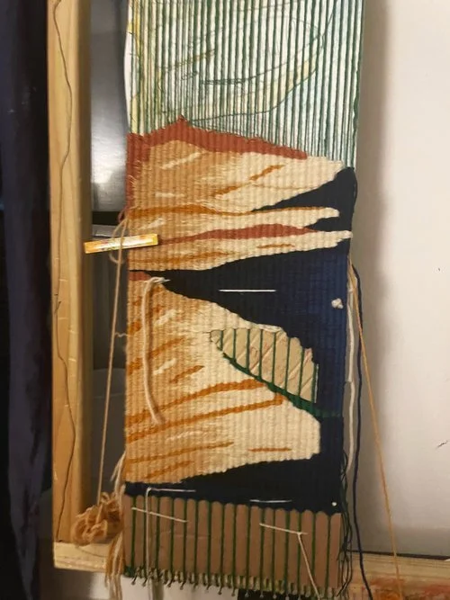 A partially woven tapestry featuring abstract patterns in black, beige, and orange on green vertical warp threads, hanging vertically with yarns and weaving tools nearby.