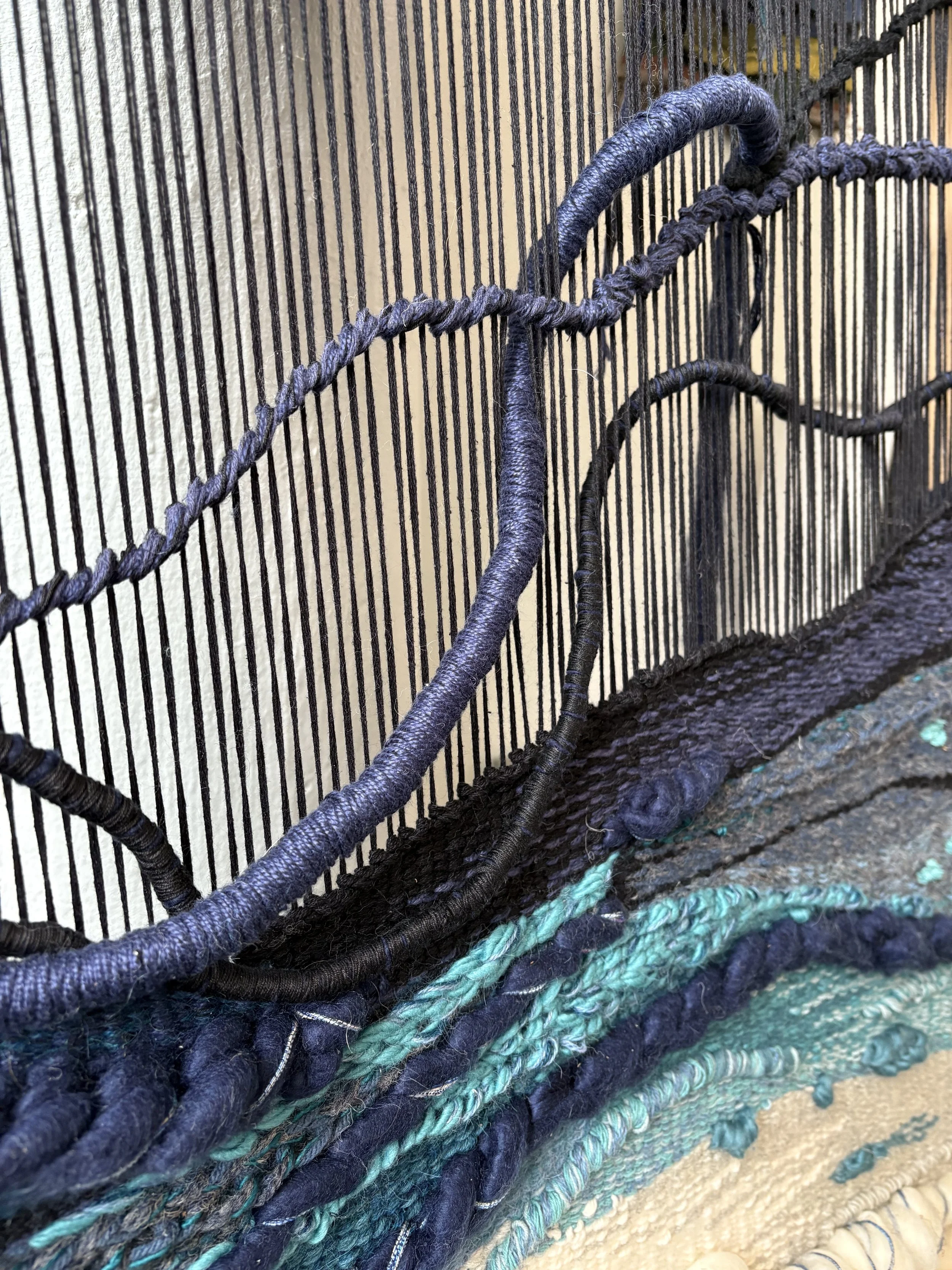 Close-up of a decorative textile piece with black, blue, and teal yarns and a black woven wire framework.