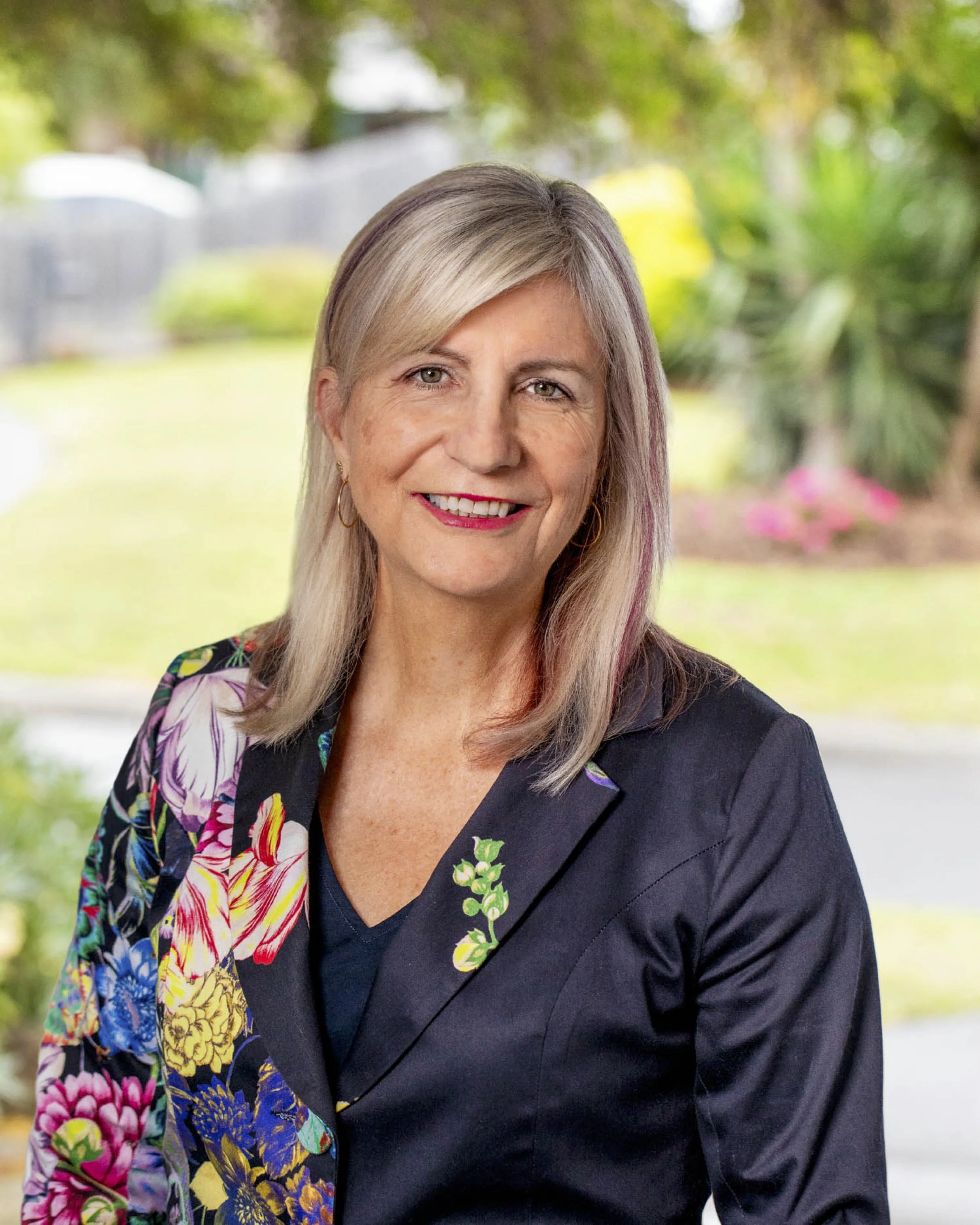 Portrait of a smiling woman with blond hair, wearing a black blazer with colorful floral patterns, set against a blurred outdoor background with greenery.