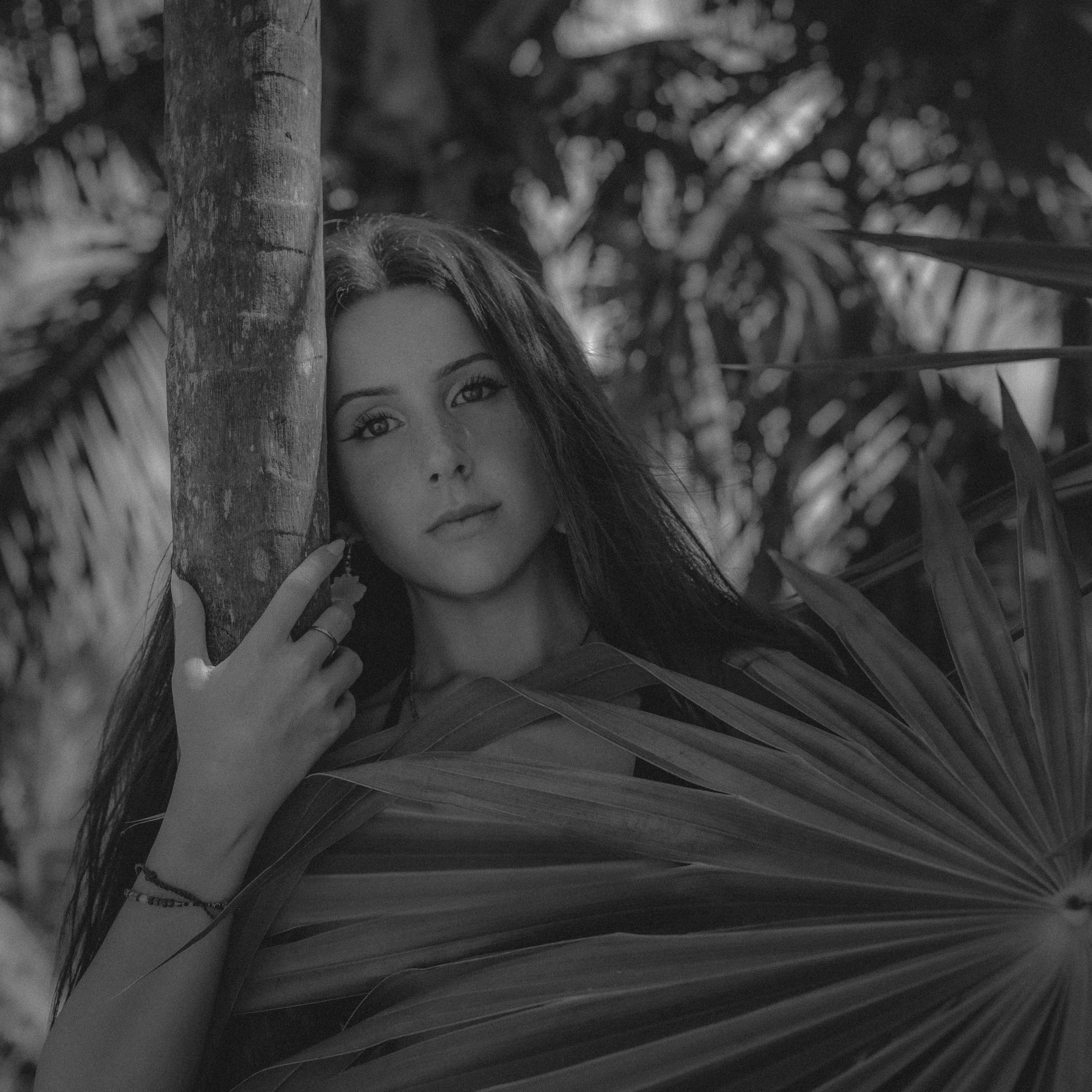 A close-up black and white photo of a woman with long hair, looking directly at the camera, with a background of blurred foliage and a tree trunk.