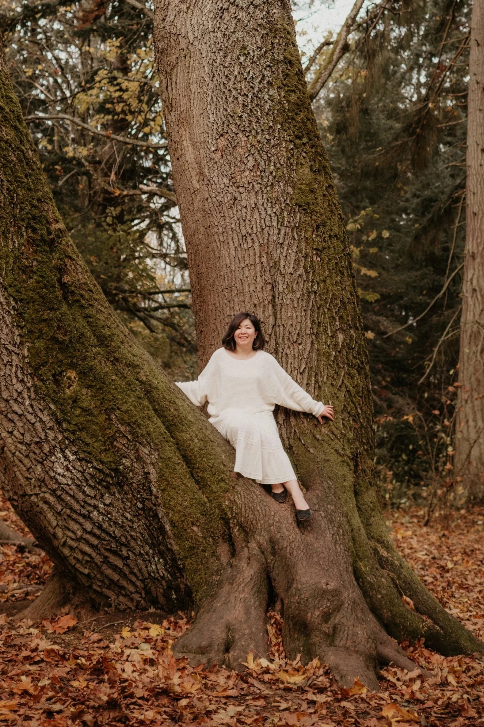 A woman wearing a white outfit sitting on a large tree branch in a forest filled with fallen leaves and tall trees.