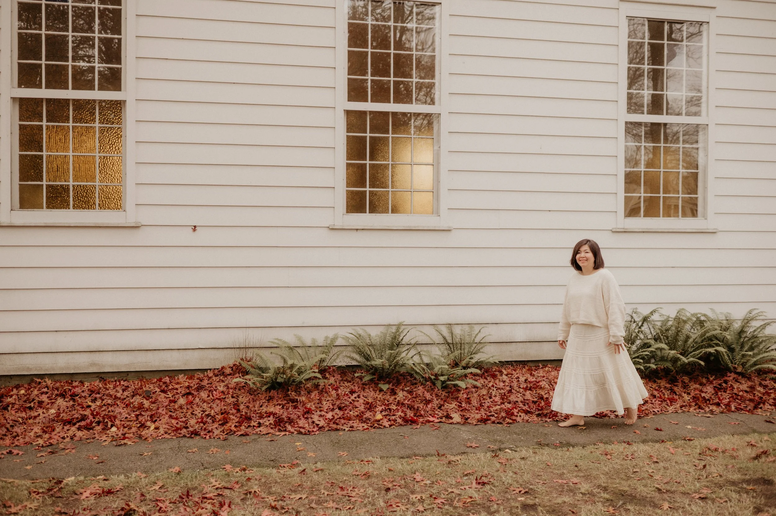 A woman in cream-colored sweater and skirt standing on sidewalk in front of a white house with four windows, autumn leaves on ground, and ferns lining the house.