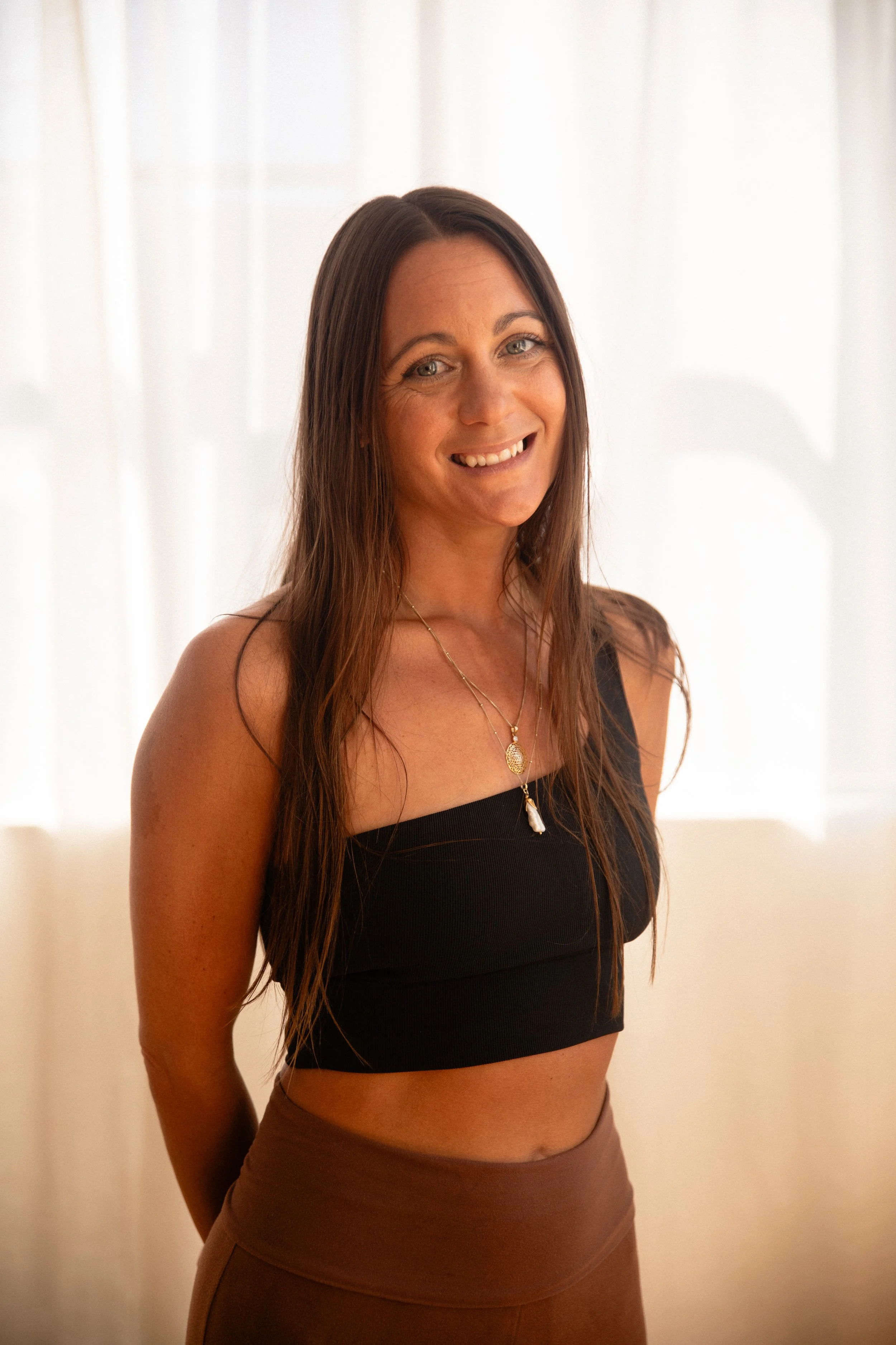 A woman with long brown hair wearing a white blouse with red and brown floral patterns, standing indoors against a beige background.