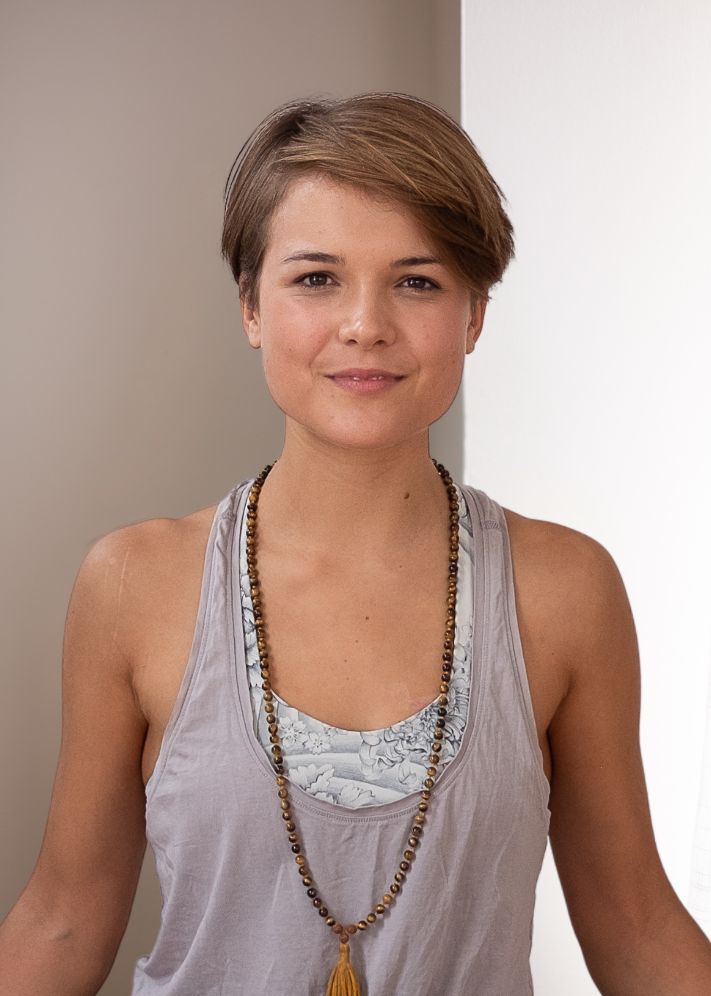 A woman with short brown hair wearing a sleeveless top and beaded necklace is smiling, standing indoors.