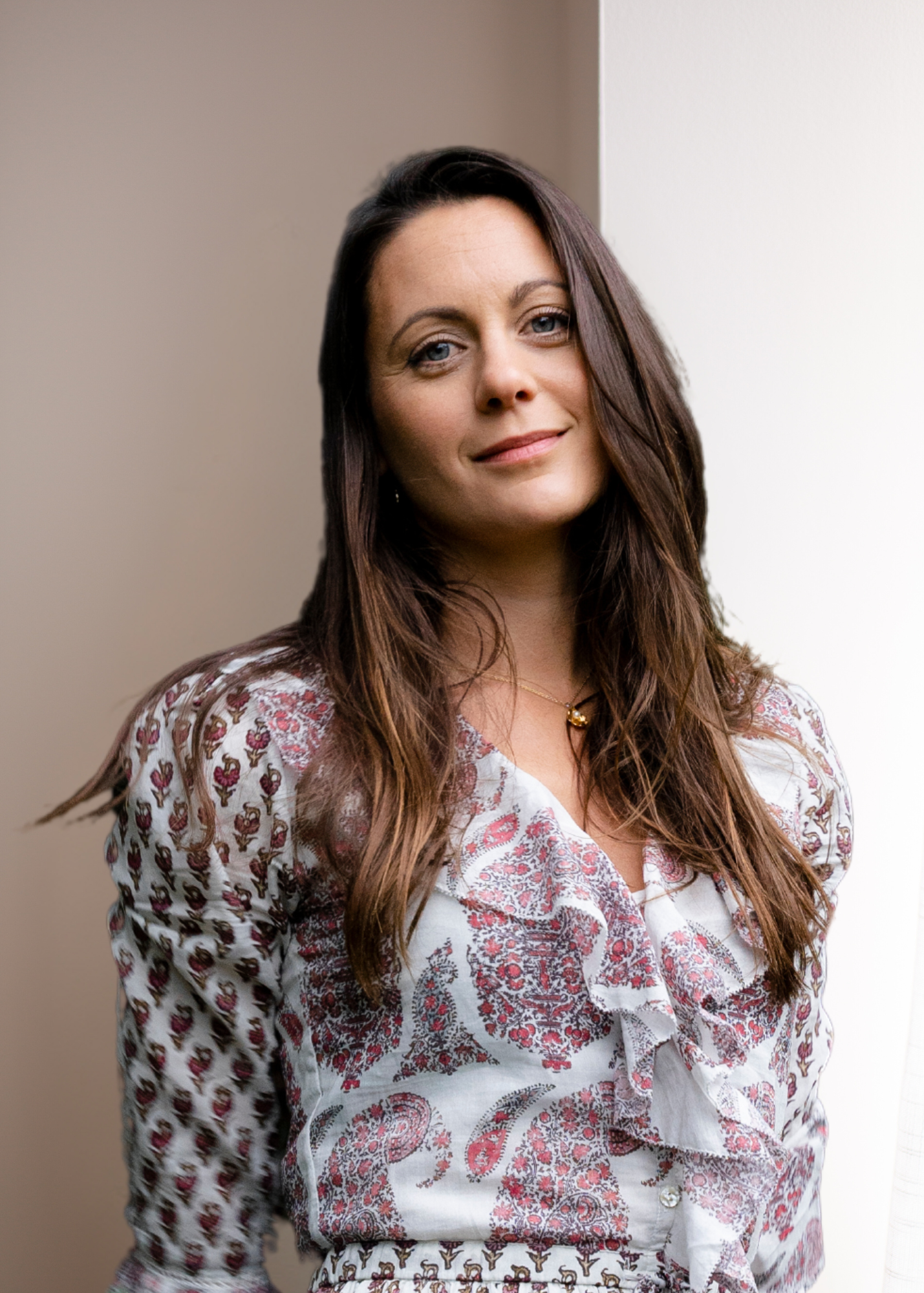 A woman with long brown hair wearing a white blouse with red and brown floral patterns, standing indoors against a beige background.