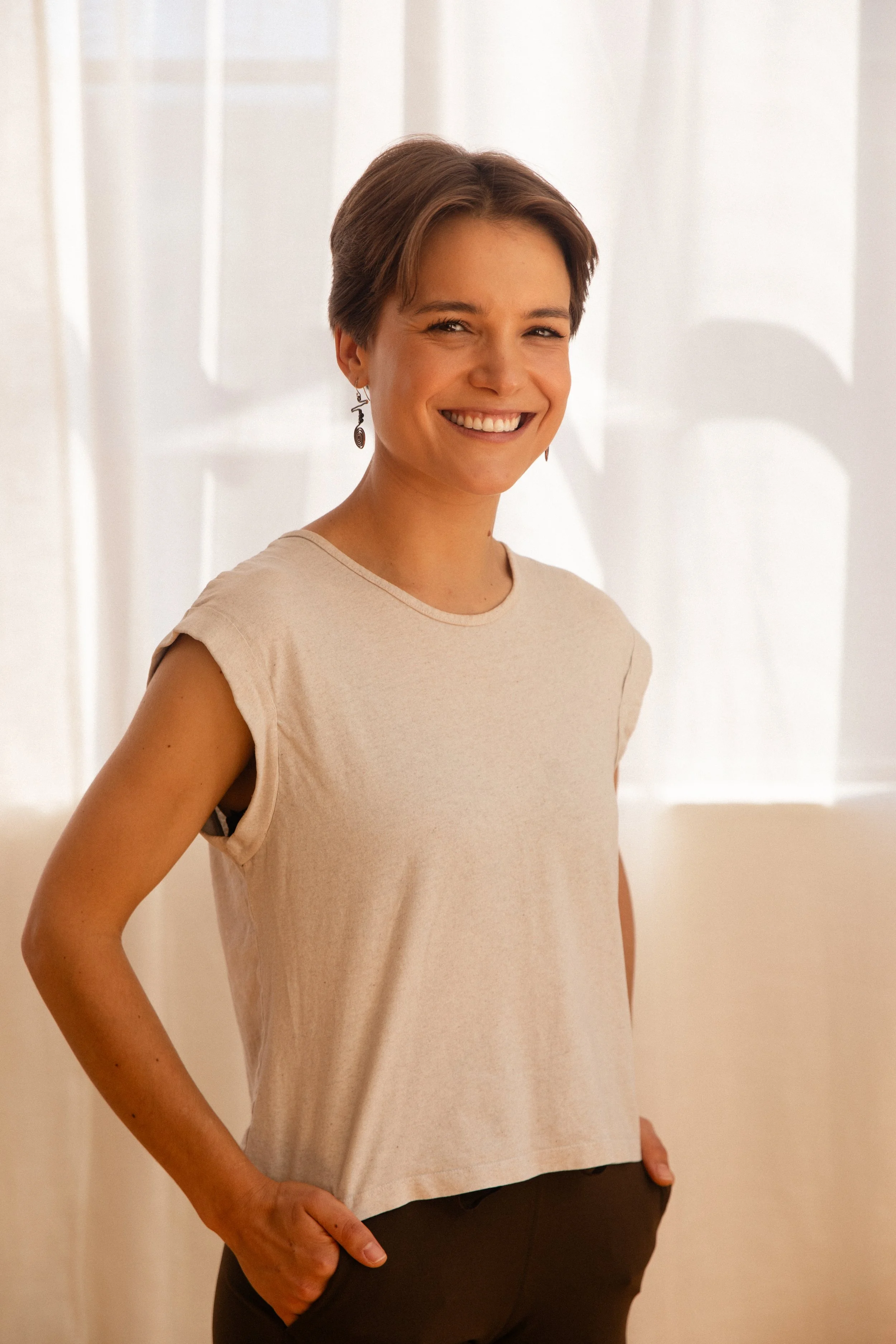 A woman with short brown hair wearing a sleeveless top and beaded necklace is smiling, standing indoors.