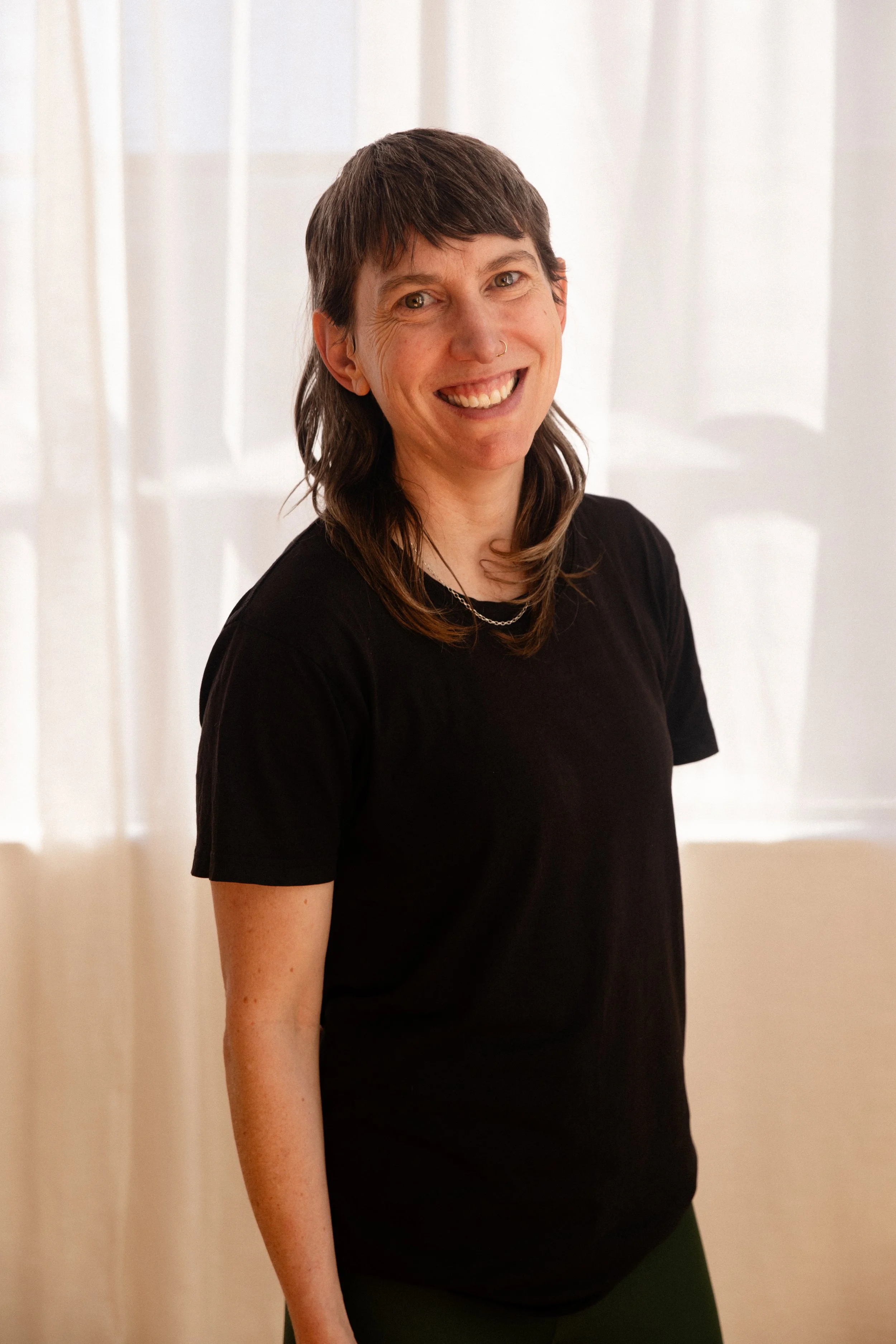 A smiling woman with short dark hair, wearing a black turtleneck, standing in front of a plain background.