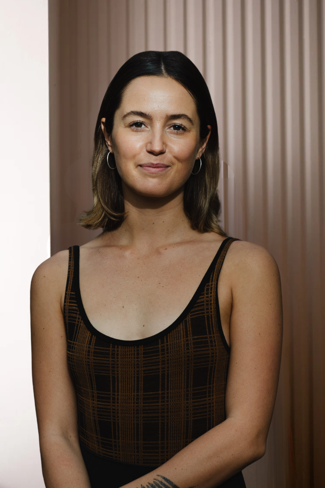 A young woman with shoulder-length brown hair, wearing a sleeveless brown and black checkered top, standing outdoors in a wooded area with dark trees in the background.