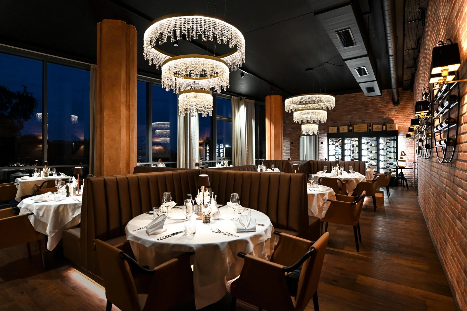 Elegantes Restaurant mit runden Tischen, weißen Tischdecken, Kerzen, modernen Stühlen, großen Kronleuchtern, Holzfußboden, brick walls, großen Fenstern mit Vorhängen, Weinschränken, und moderner Beleuchtung.