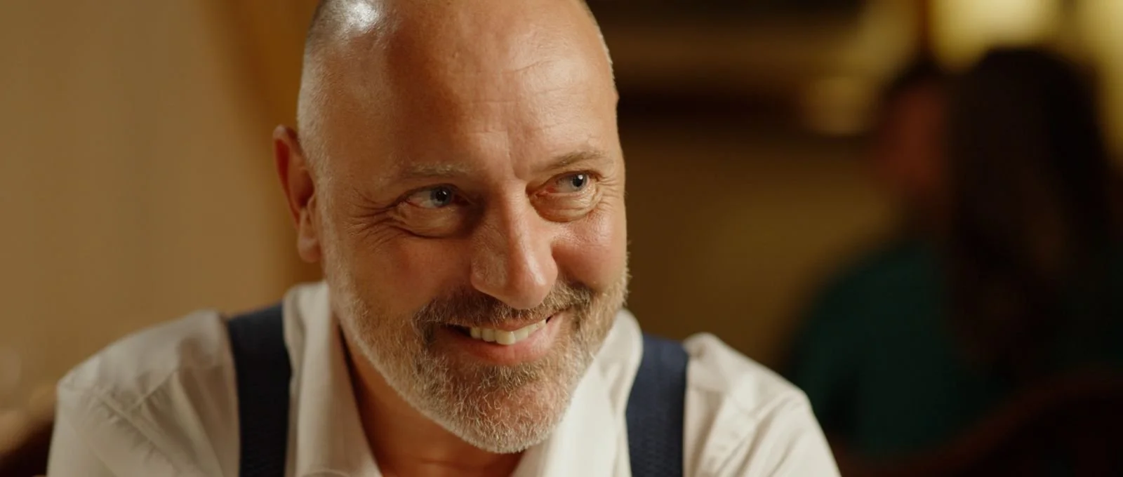 A smiling man with a bald head and white shirt in a warm, dimly lit setting.