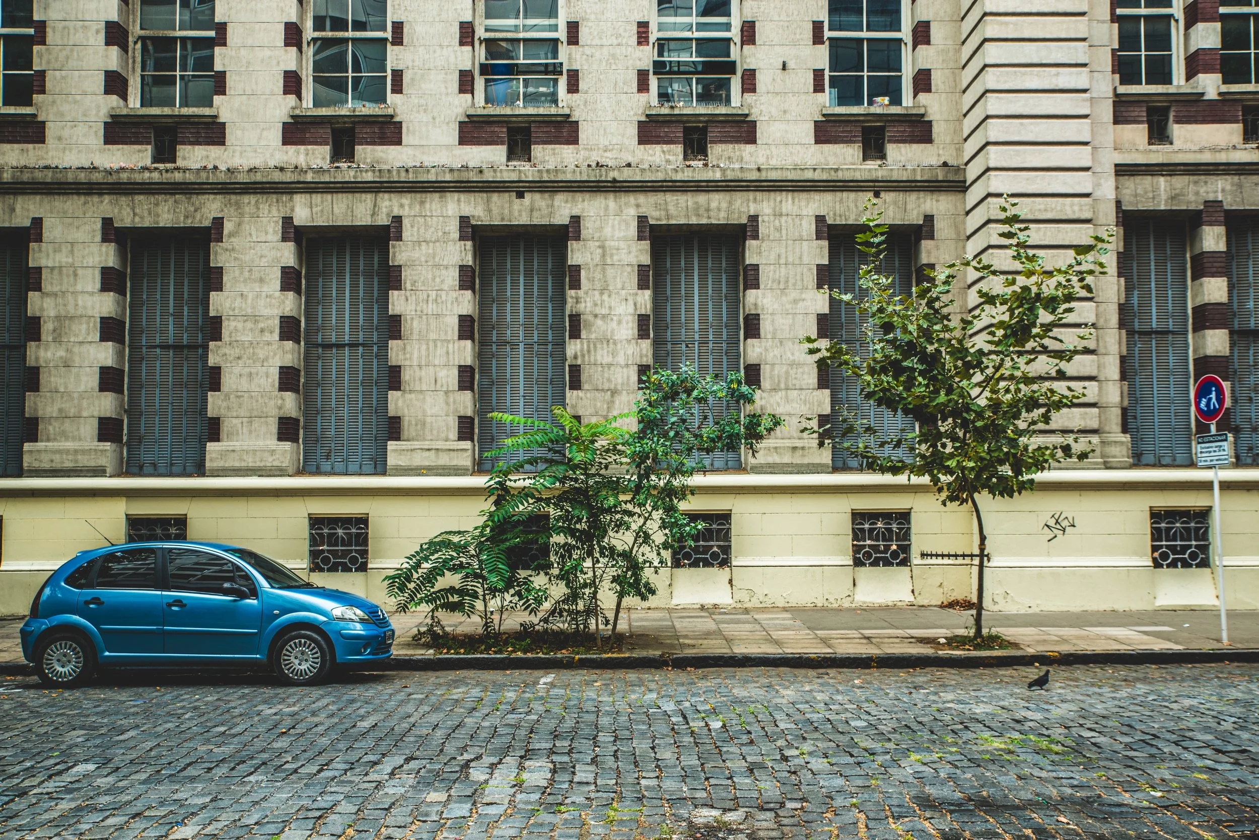 Edificio de arquitectura clásica del siglo XIX con un auto moderno estacionado frente a él, representando la tensión entre lo que cargamos del pasado y la vida cotidiana que sigue.