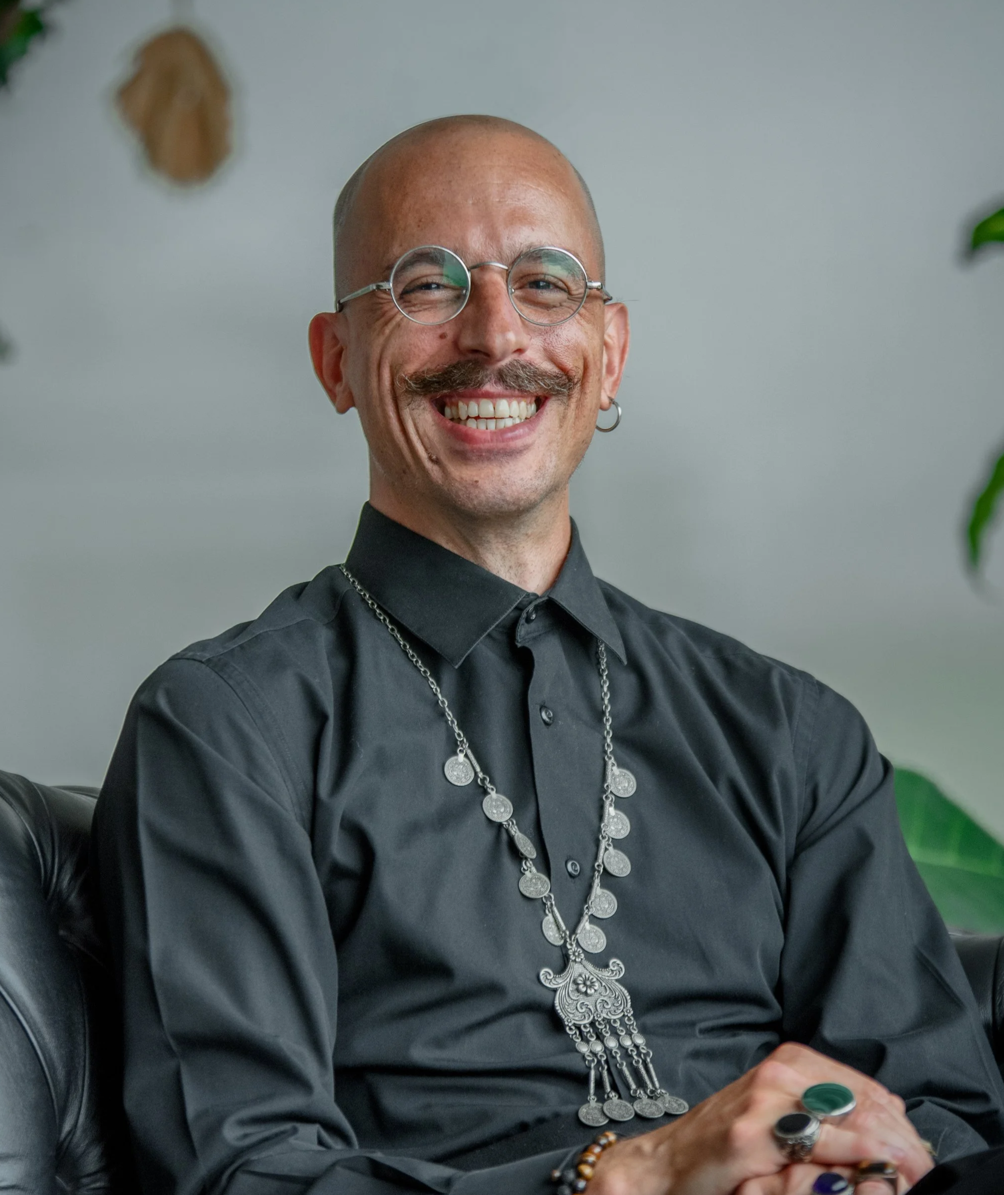 A smiling person with a bald head, round glasses, a mustache, and earrings, dressed in a black shirt and silver jewelry, sitting on a black leather chair against a grey background with green plants on the sides.