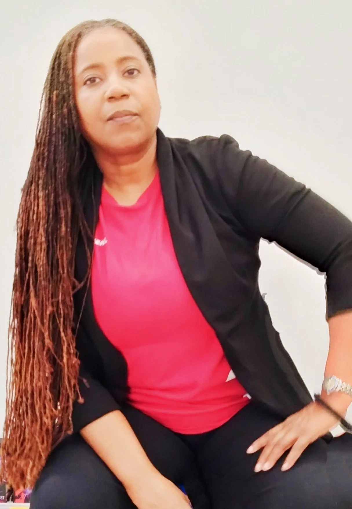 woman with long braided hair sitting against a white wall, wearing a red shirt and black cardigan.