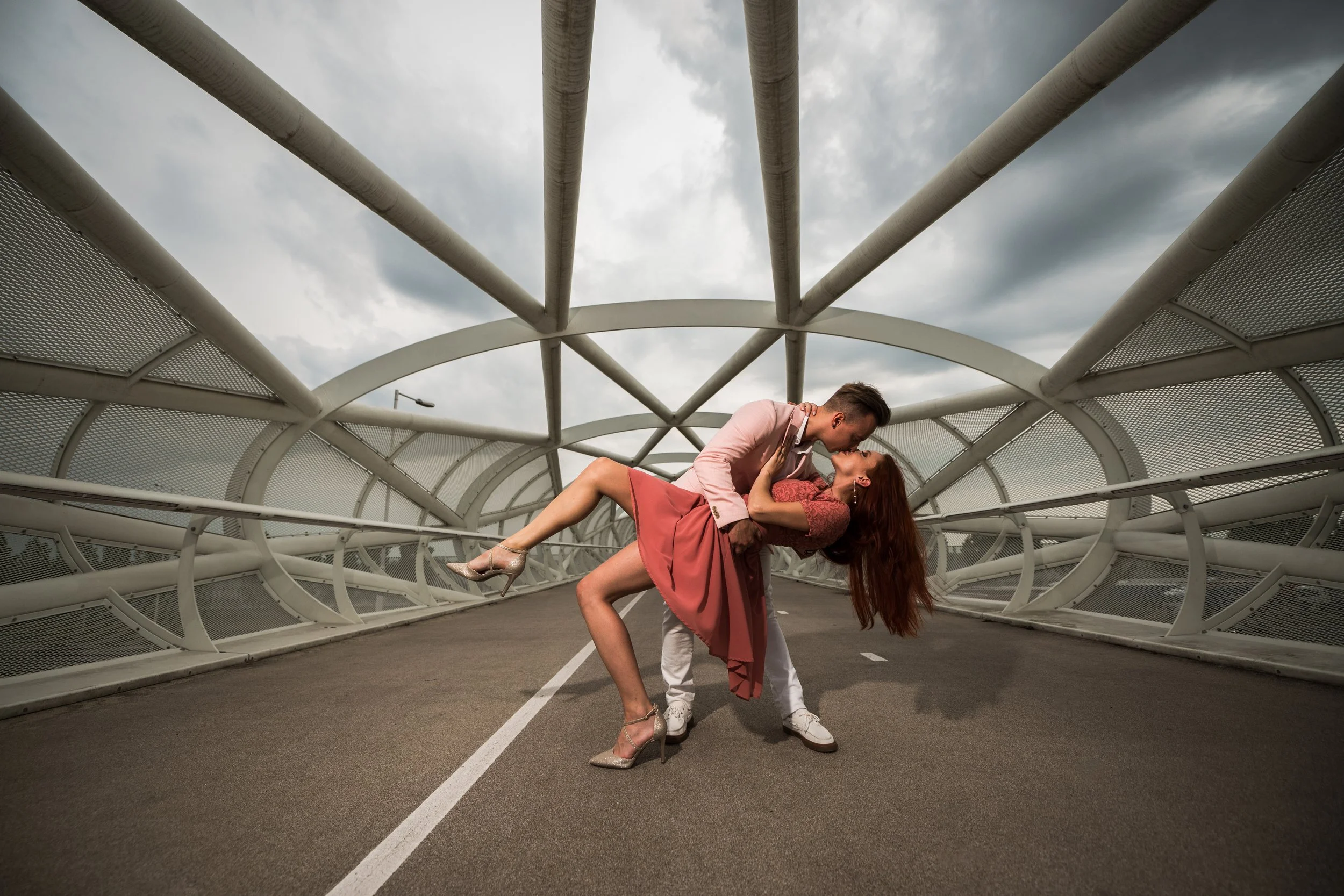 A man and woman dance on a modern bridge with a geometric metal structure under a cloudy sky. The man is in a light pink jacket and the woman wears a pink dress.