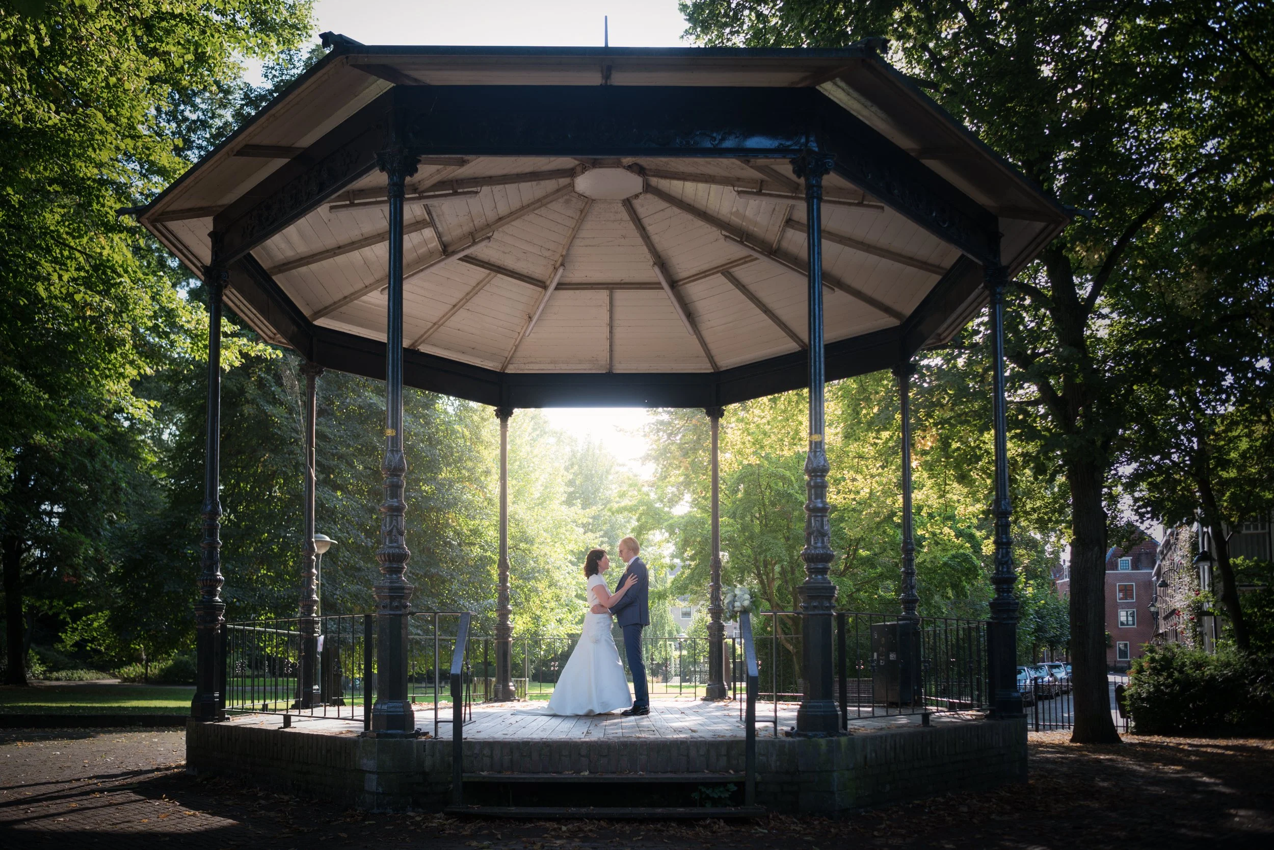 A bride and groom dancing in a gazebo in a park surrounded by green trees, with sunlight shining through.