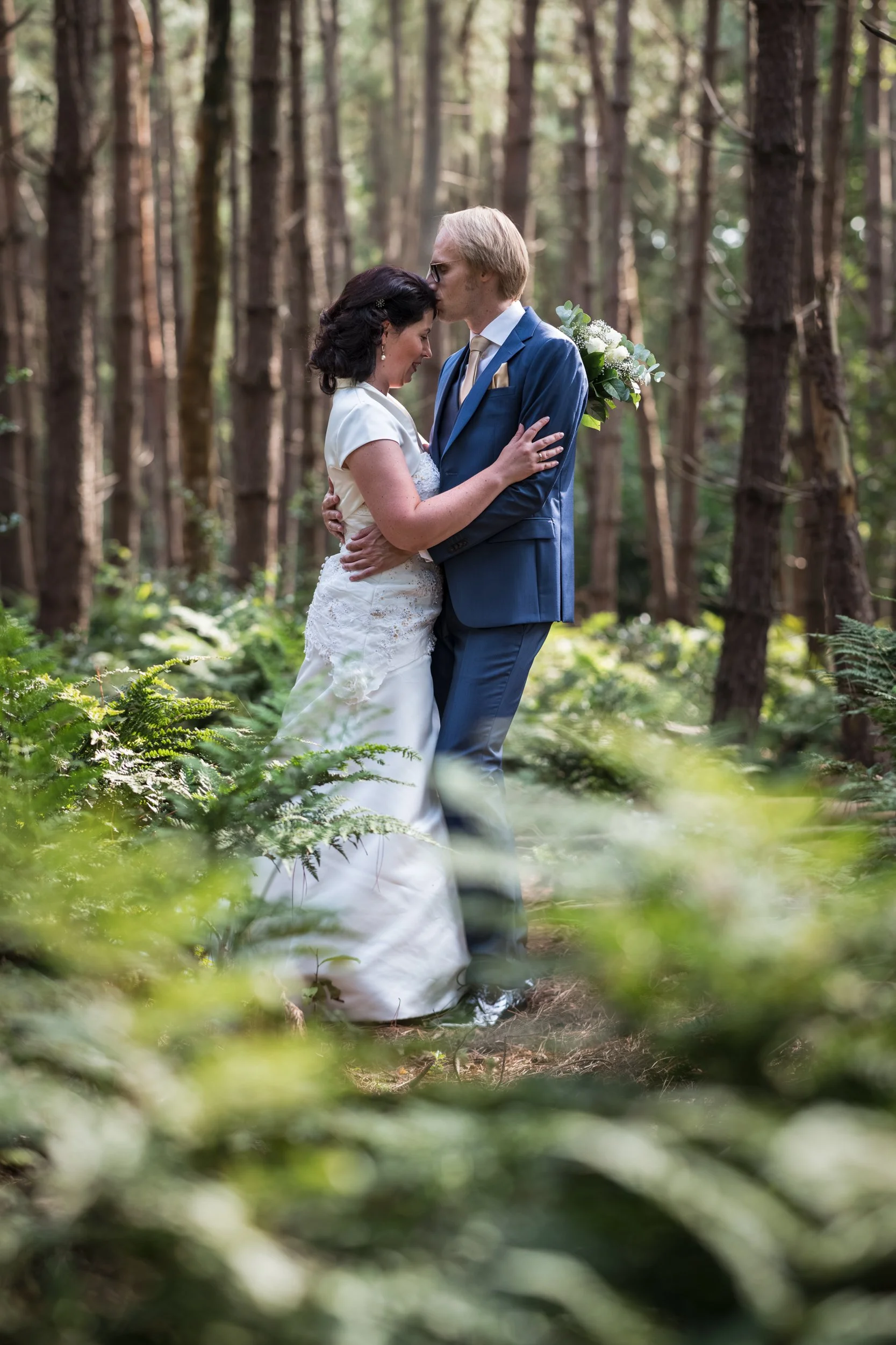A newlywed couple embraces in a wooded forest, with the groom kissing the bride's forehead, surrounded by tall trees and green foliage.