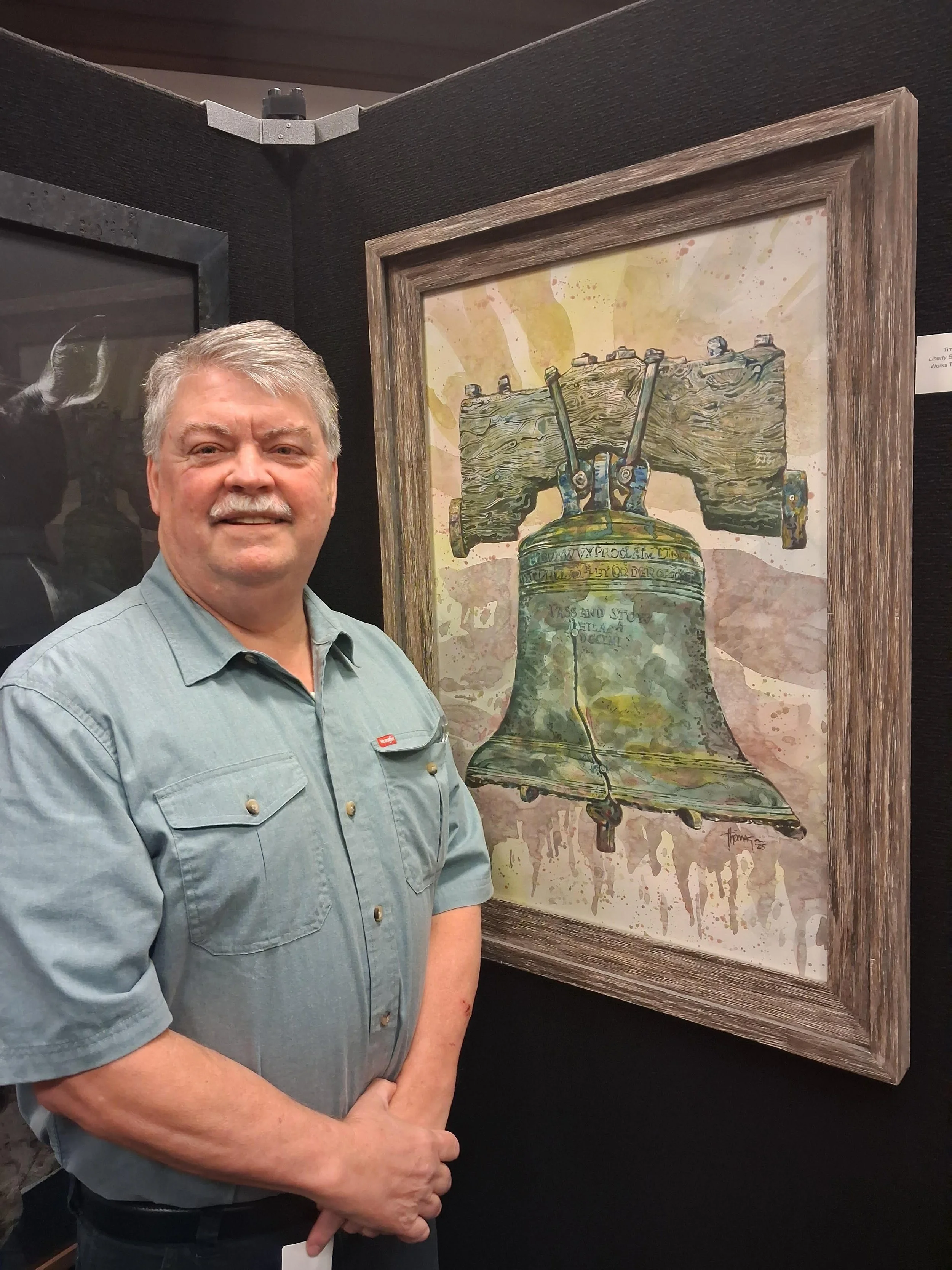 A smiling man with gray hair and a mustache, wearing a light blue shirt, standing next to a framed painting of the Liberty Bell in an art gallery.