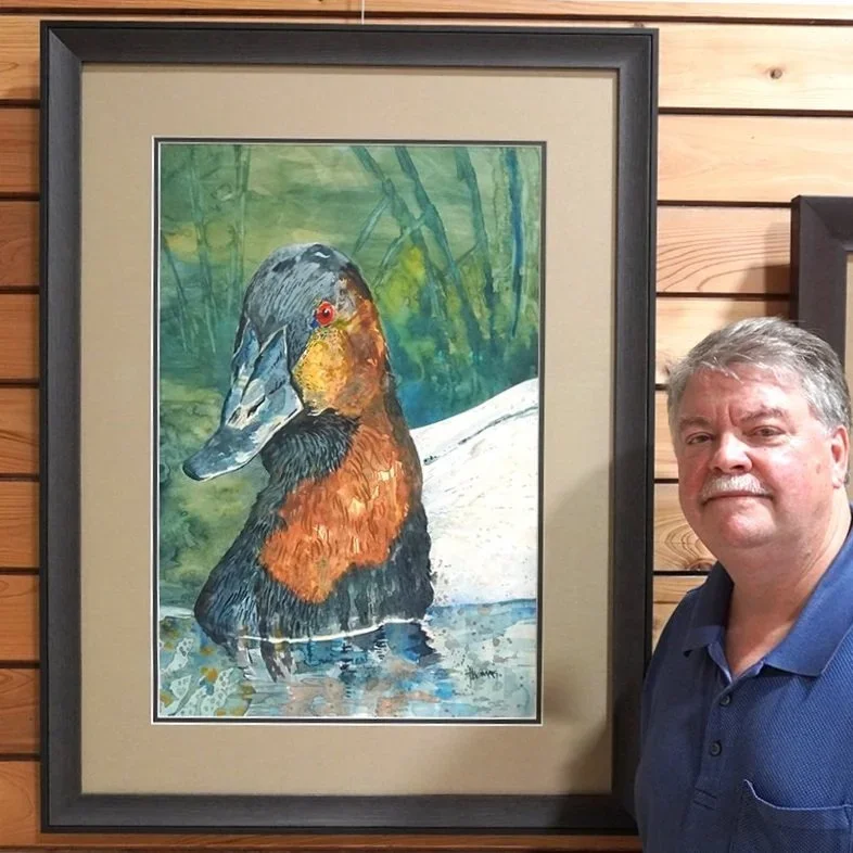 Portrait of a man standing next to a framed watercolor painting of a duck, with a wooden wall background.