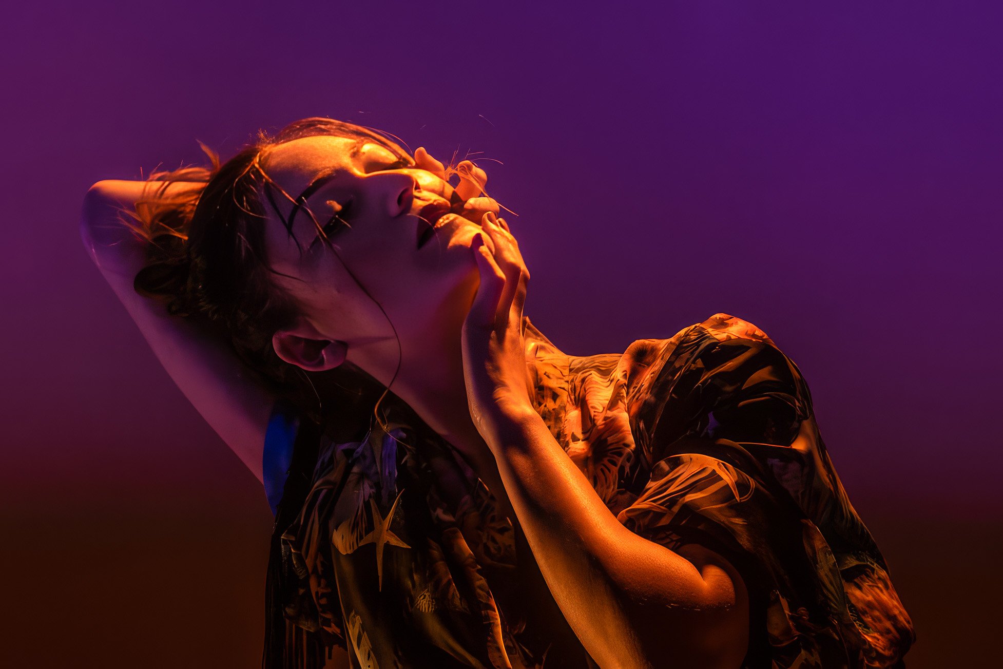 A woman with wet hair poses with her hand on her neck and her eyes closed, illuminated by purple and orange lighting, wearing a patterned shirt.