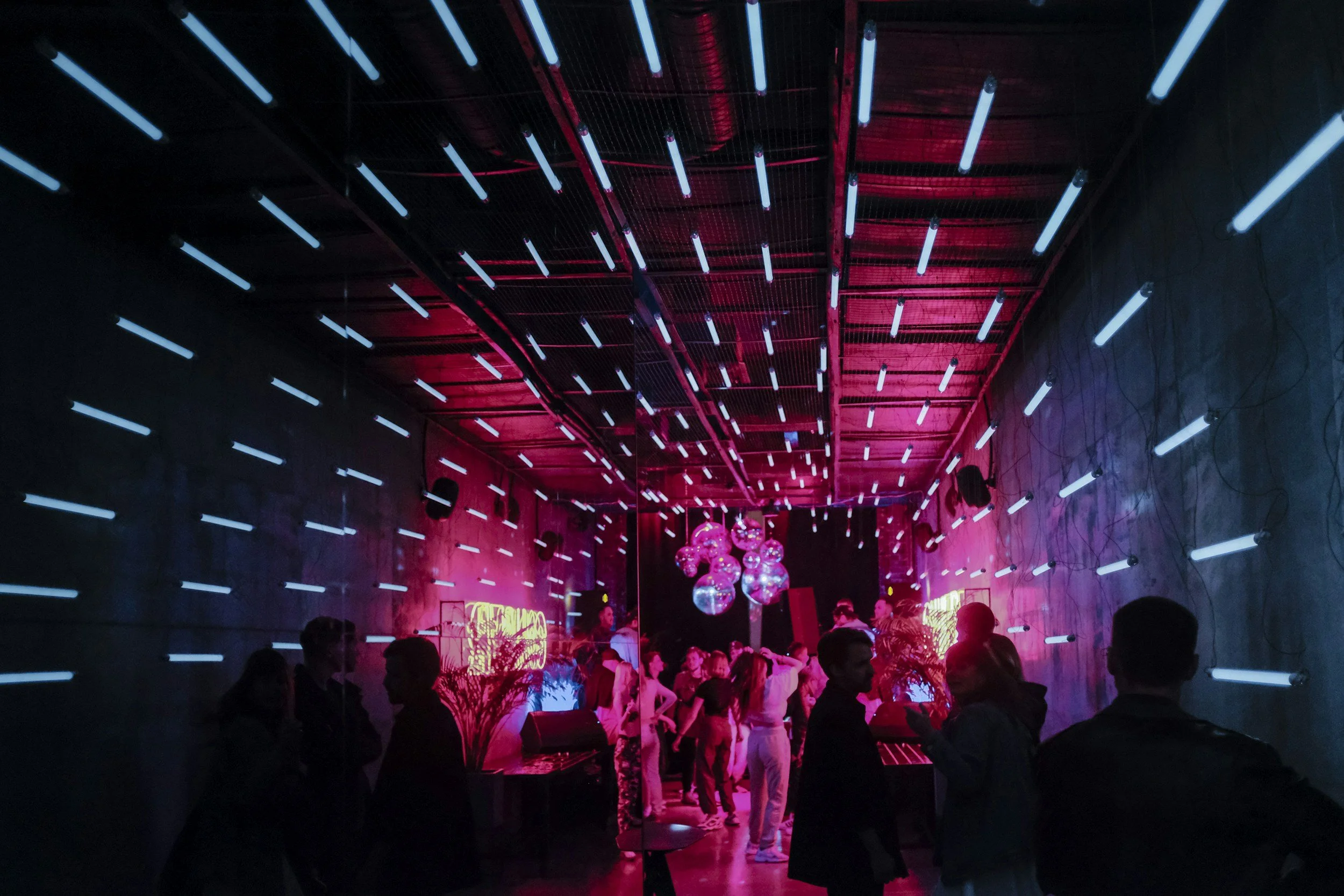 Guests at a small party turned disco, surrounded by vibrant lighting in a dark atmosphere. Tube lights on the walls and ceiling create a tunnel effect, adding to the lively and energetic vibe of the event