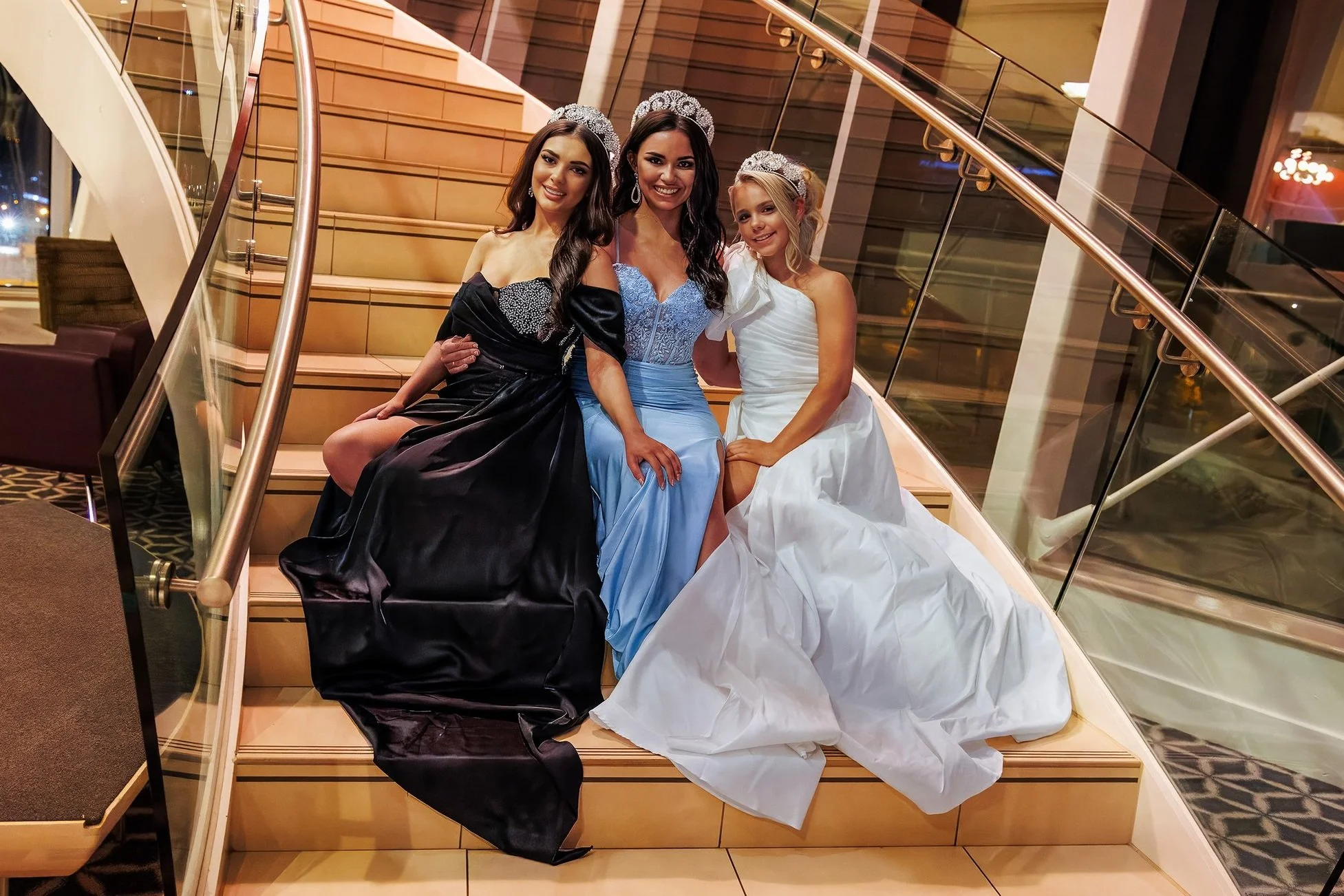 Three beauty queens in elegant evening gowns, smiling and posing on a grand spiral staircase with a glass balustrade, exuding glamour and sophistication