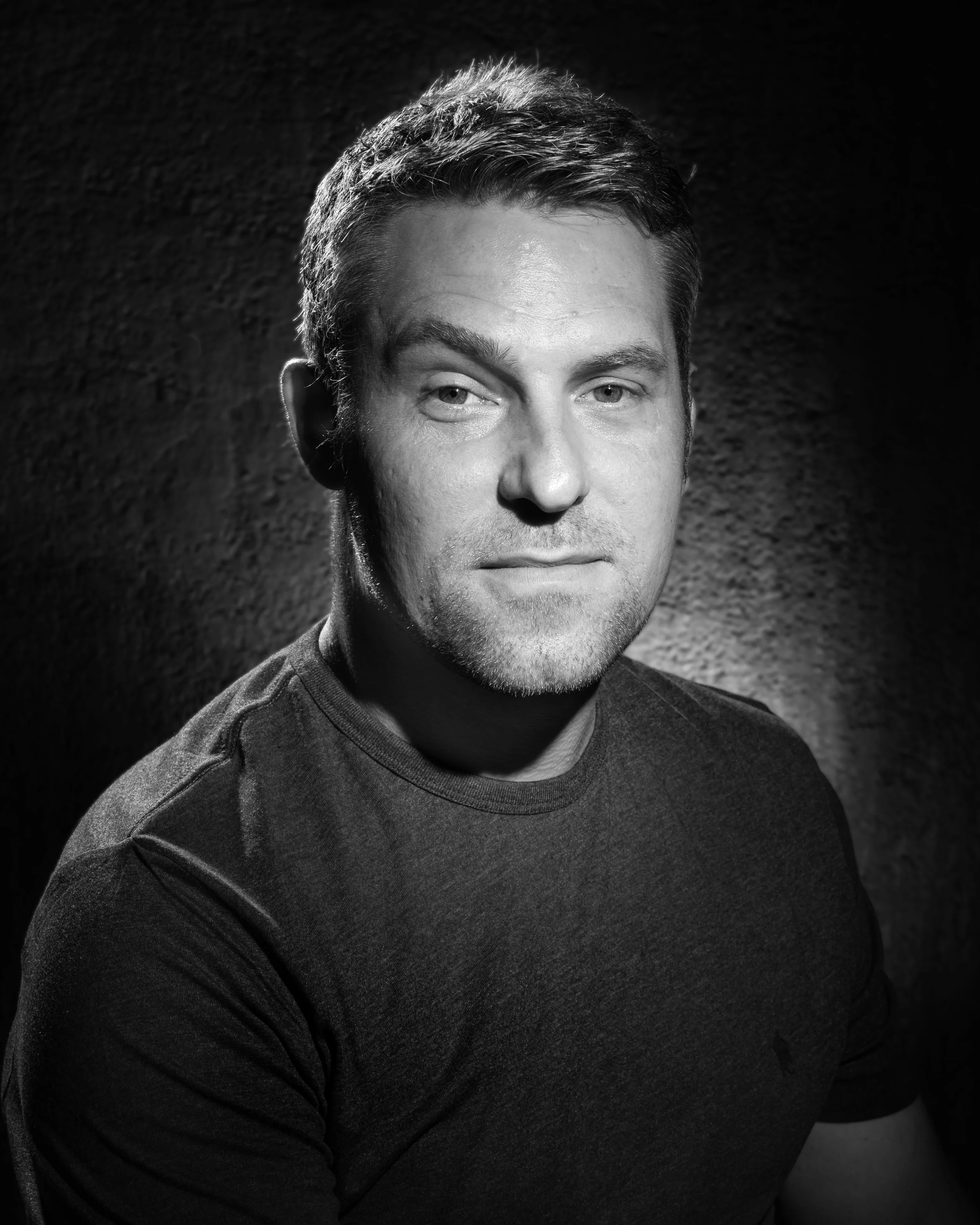 Studio portrait of actor Anthony Hayes in dramatic low-key black and white lighting, facing forward with a neutral expression.