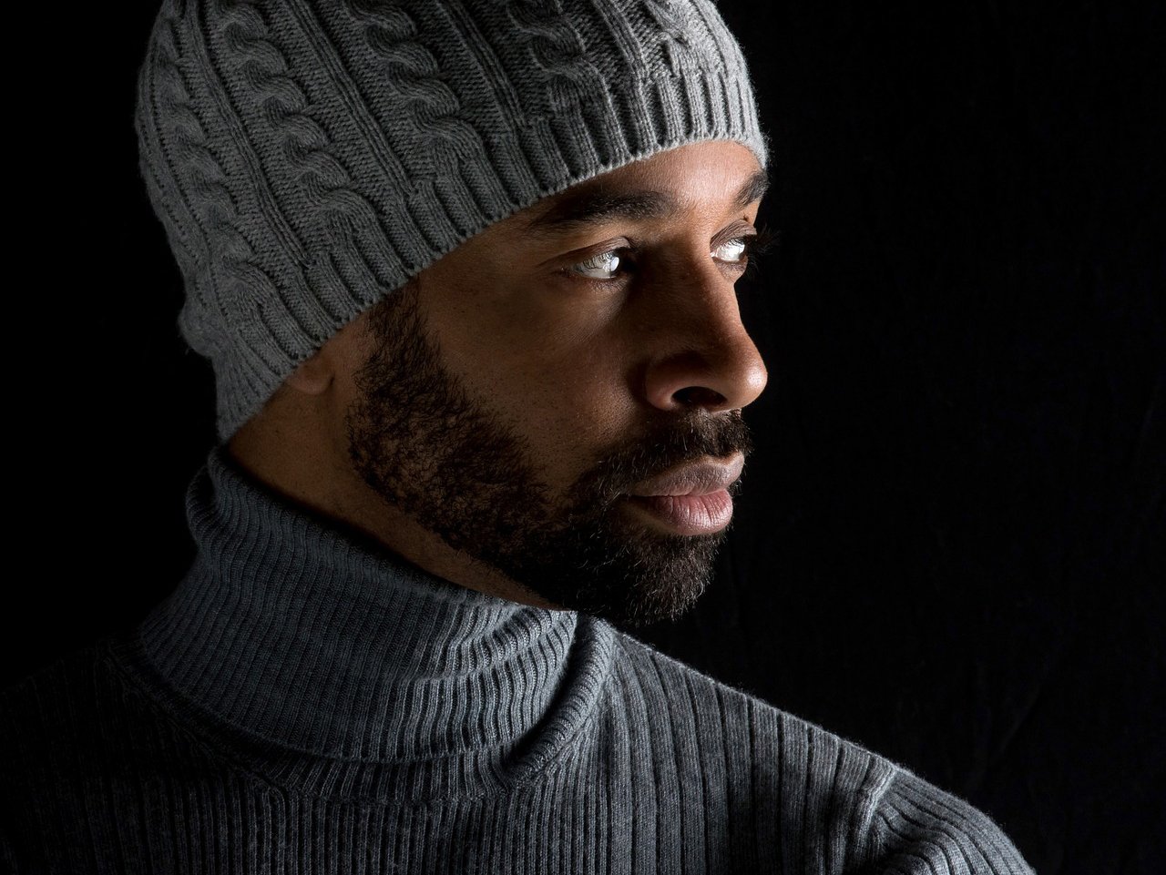 Low-key promotional headshot of a young black man wearing a grey roll neck top and a grey woolen hat, set against a neutral background, exuding a relaxed and stylish look