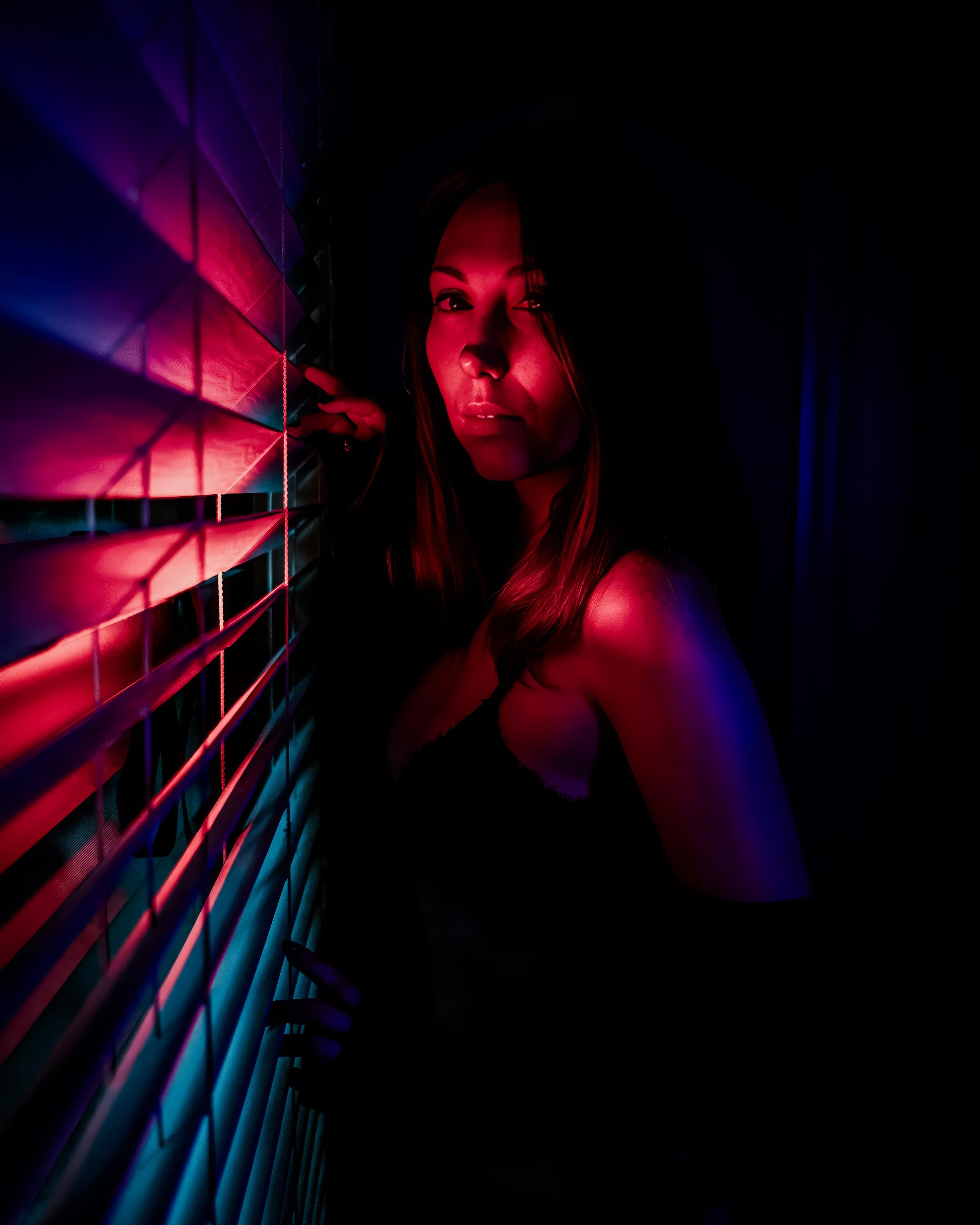 A woman in a dark room with pink and purple lighting, standing beside window blinds, looking at the camera.