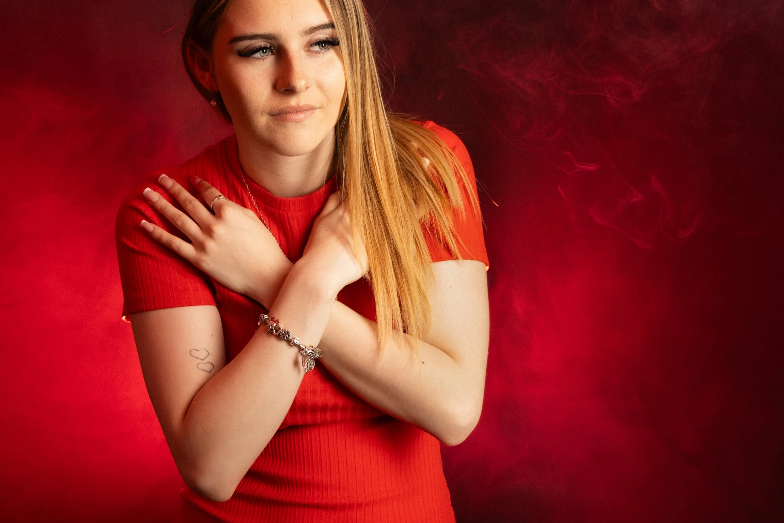 A young woman with long red hair, wearing a red shirt, poses with her arms crossed over her chest against a red background with swirling smoke.