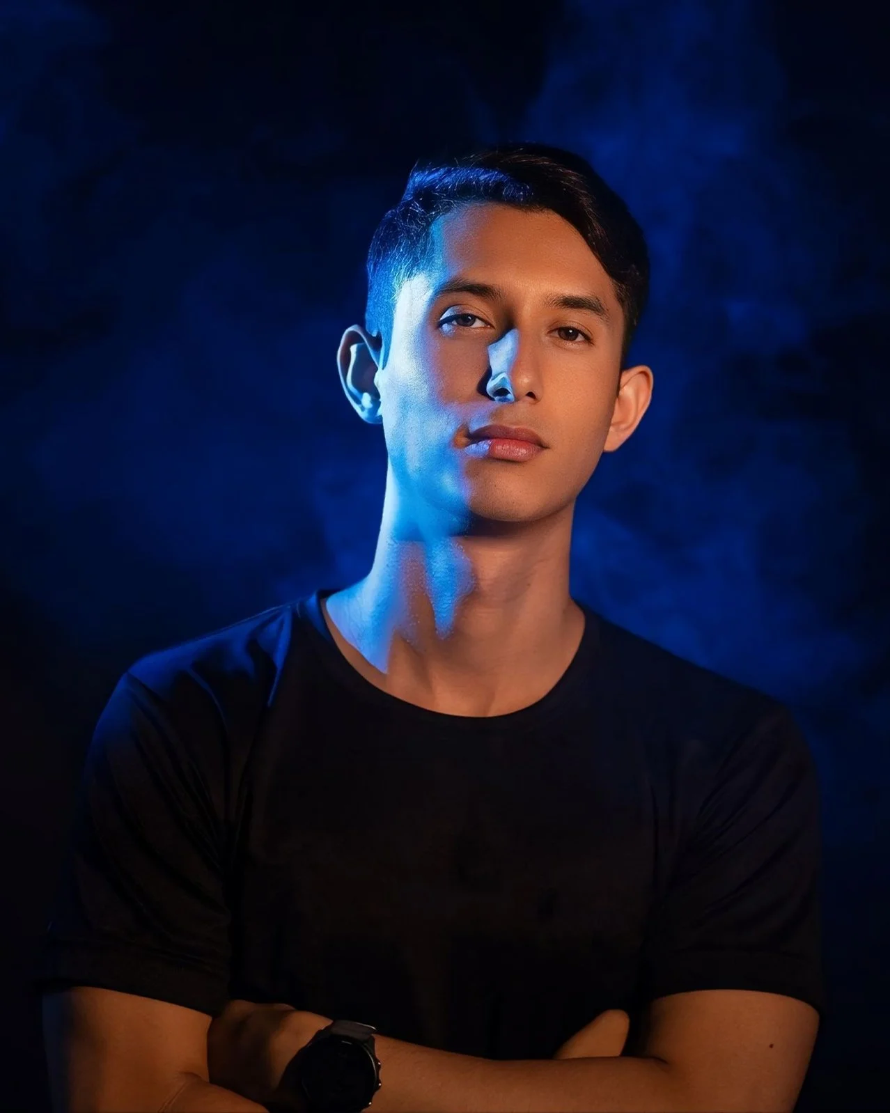 A young man with short dark hair and tan skin stands with crossed arms against a dark background with blue lighting, looking at the camera with a neutral expression.