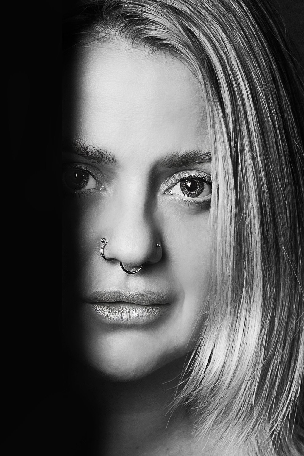 Close-up black-and-white portrait of a young woman with light-colored hair, piercings on her nose and lip, and light makeup. She gazes slightly to the side with intense eyes.