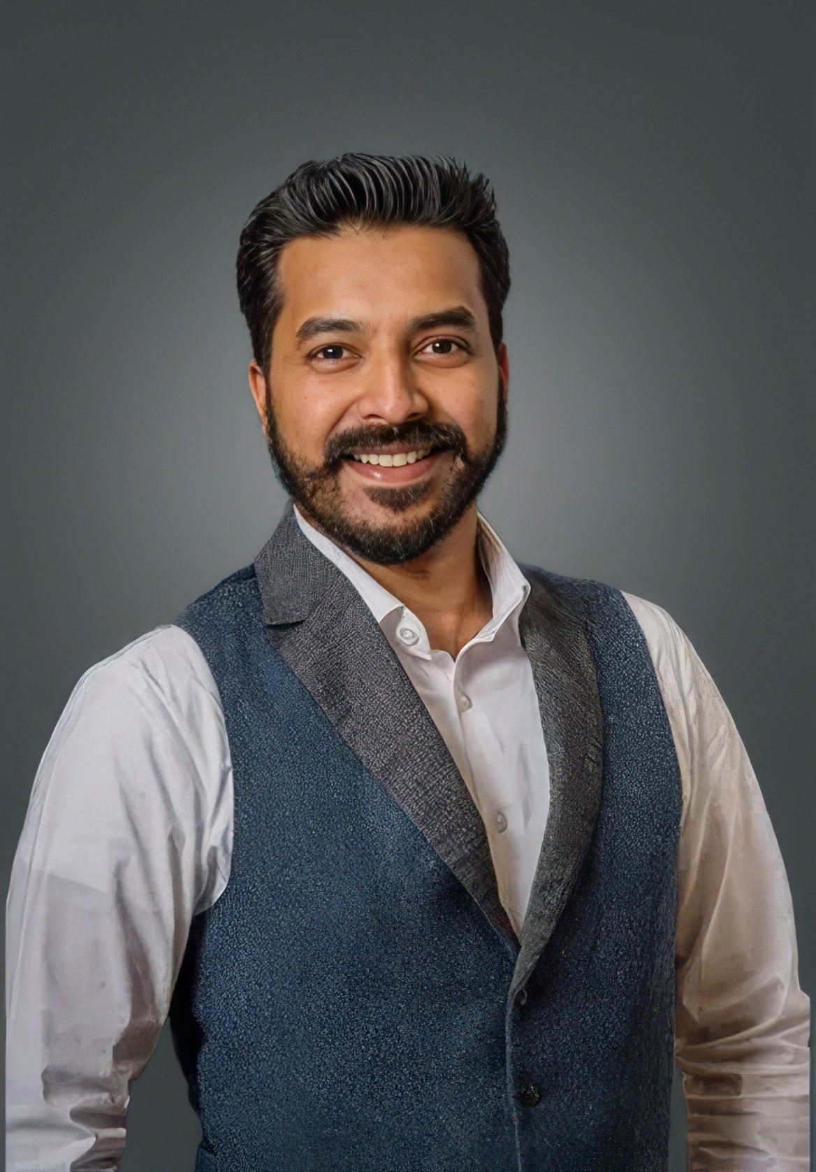 Smiling man in a white shirt and blue vest against a gray background.