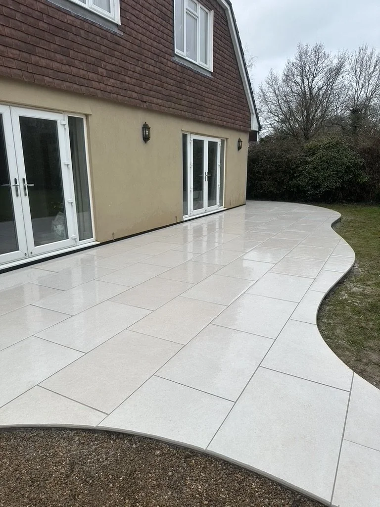 Nowo wyłożony taras z jasnymi płytami, przylegający do domu z wieloma szklanymi drzwiami, sąsiadujący z trawnikiem i częścią ogrodową.
