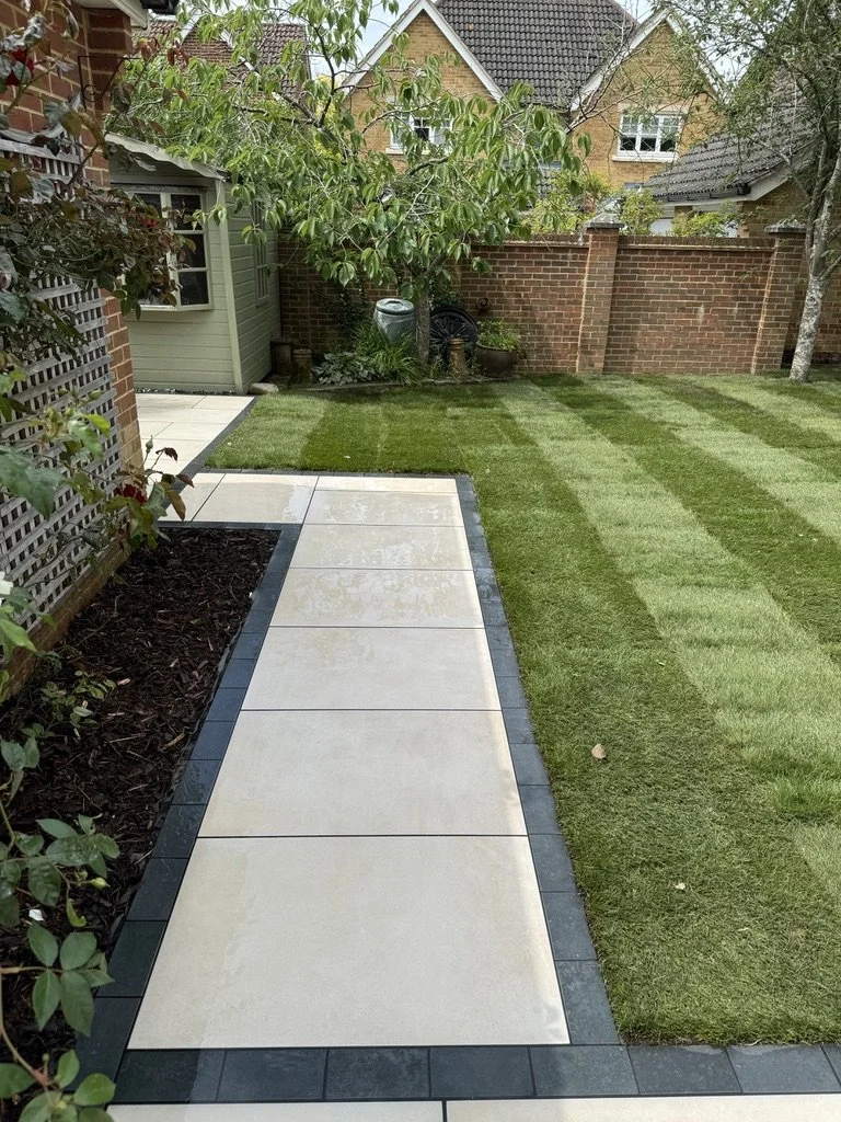 Ogród z kamienną ścieżką, zielonym trawnikiem, małym domkiem gospodarczym, ceglanym murem oraz drzewem o gęstych liściach.
