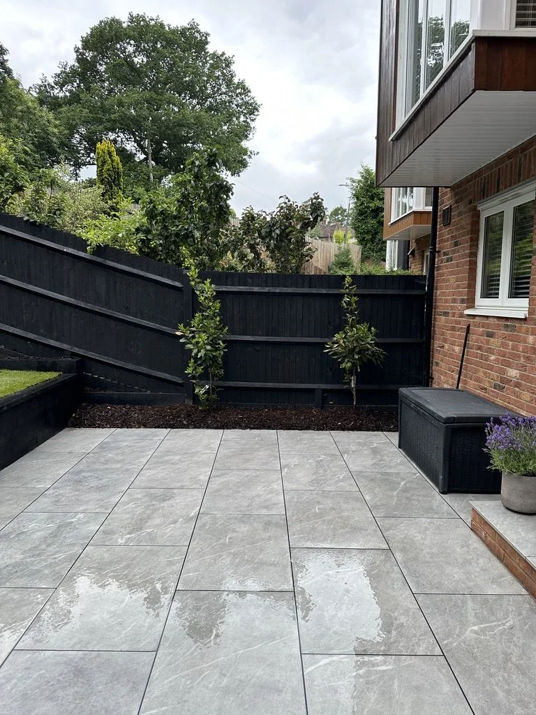 Nowoczesny taras w ogrodzie, wyłożony eleganckimi szarymi kamiennymi płytami, otoczony ciemnym drewnianym ogrodzeniem, z elewacją z cegły, uzupełniony doniczką z lawendą, która wprowadza odrobinę naturalnego koloru i zapachu