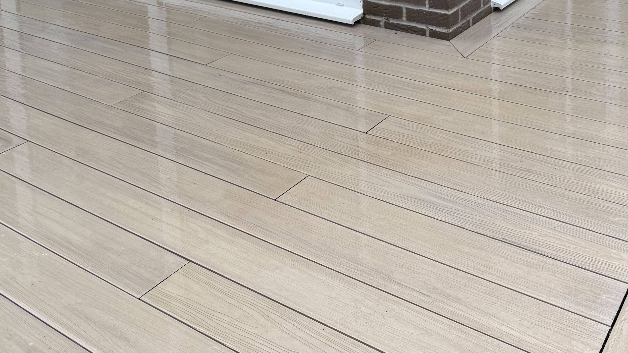 Zbliżenie na błyszczącą, beżową podłogę drewnianą z widocznymi deskami, w tle ceglany mur.
