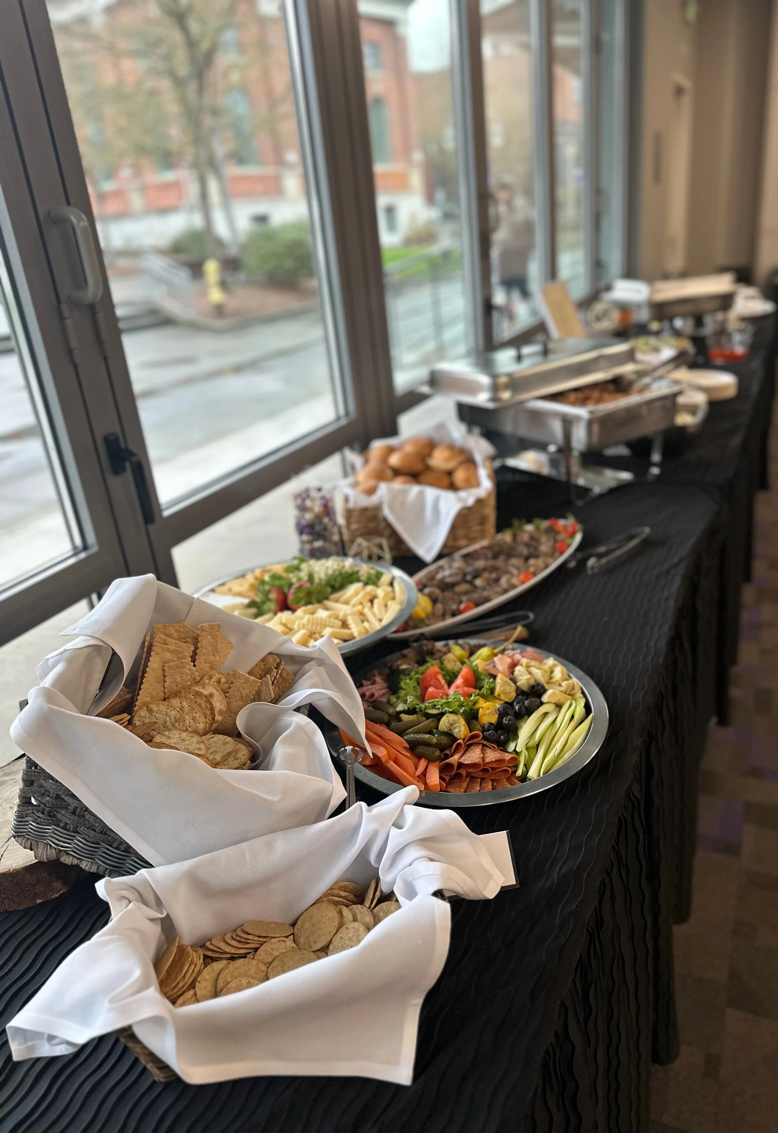 Buffet table with various dishes including crackers, vegetables, cheese, salads, and meat, set by a window in a dining area.