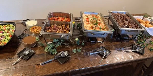 Buffet of garden salad, roasted potatoes, roasted carrots, chicken in a caprese sauce, beef, and rolls on a wooden table decorated with vines.