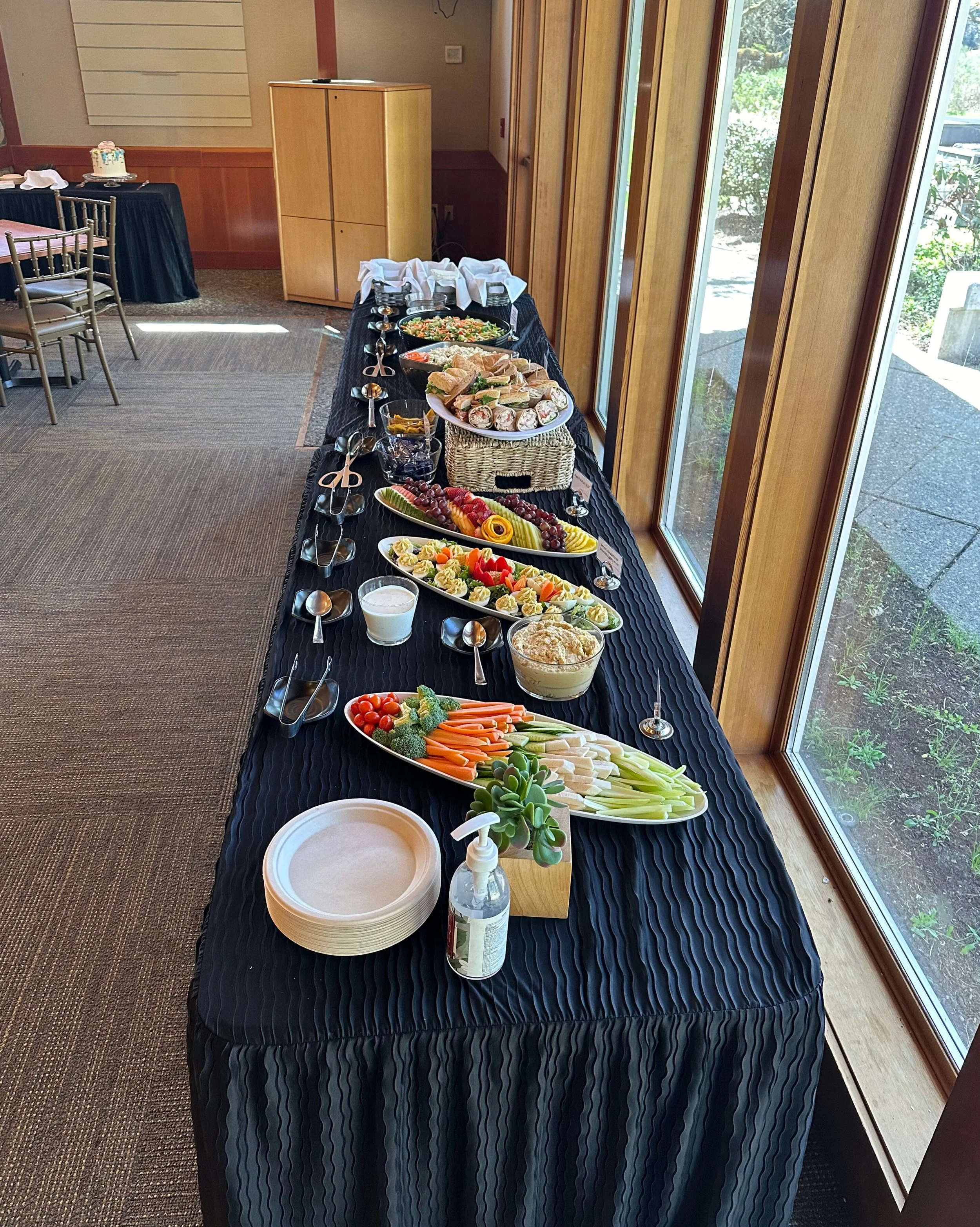 Buffet table with various appetizers, vegetables, and finger foods set near large windows in a decorated room for a gathering or event.