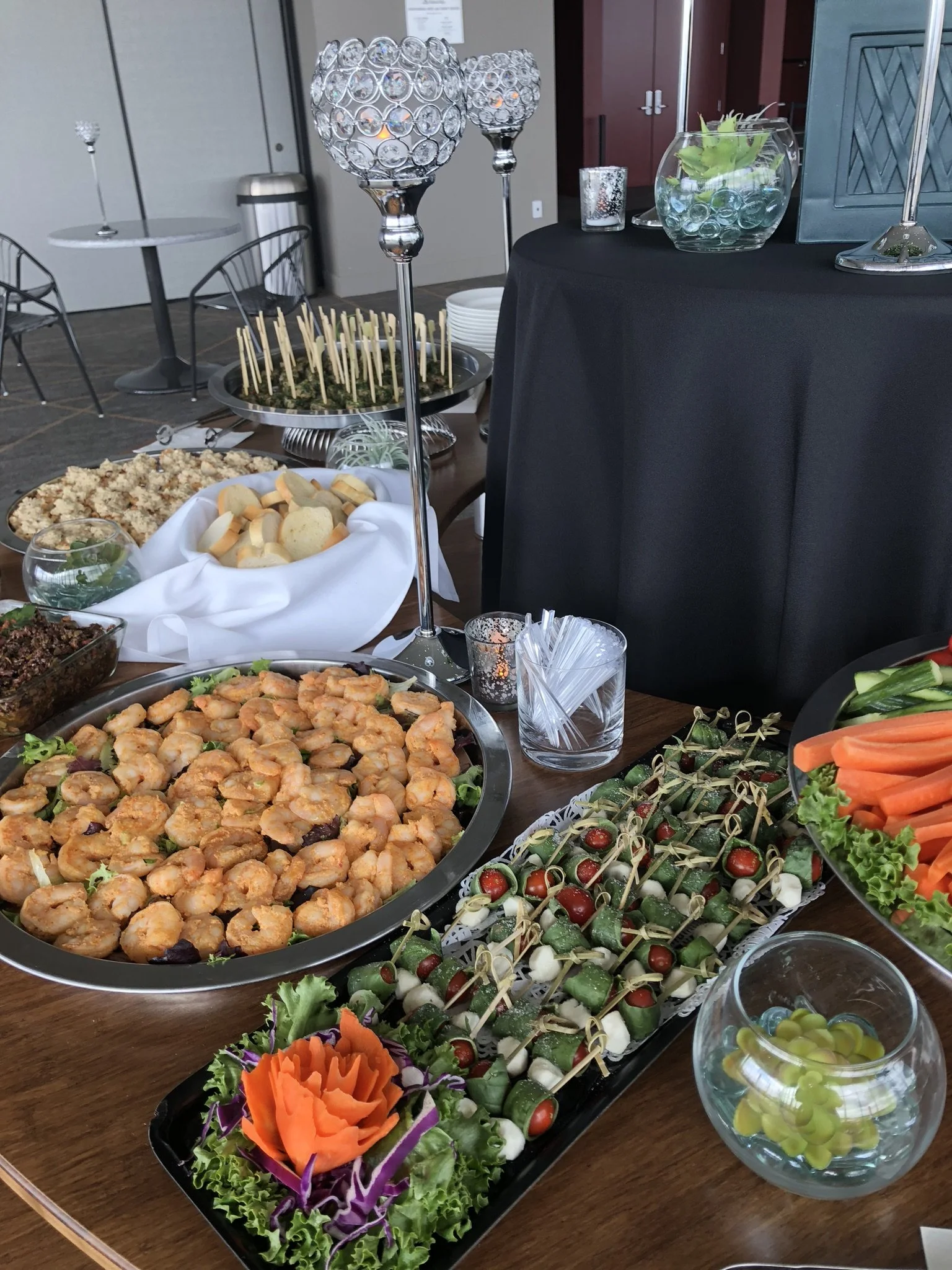 A table with various appetizer dishes including shrimp, cucumber and cherry tomato skewers, carrot sticks, and cheese, decorated with candles and glass bowls of grapes and vegetables, set up for a celebration or event.