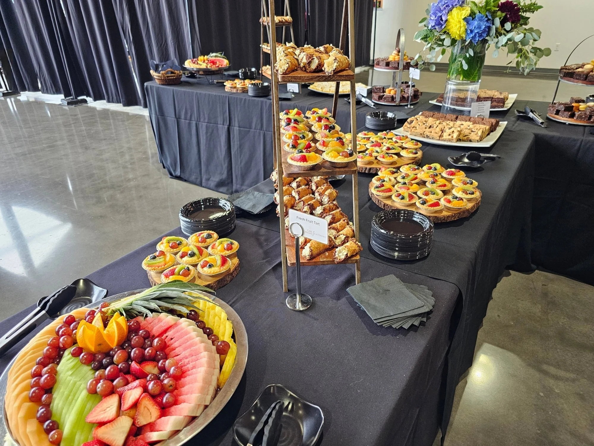 Black buffet tables filled with trays of fruit tarts, brownies, blondies, fruit, and other desserts.