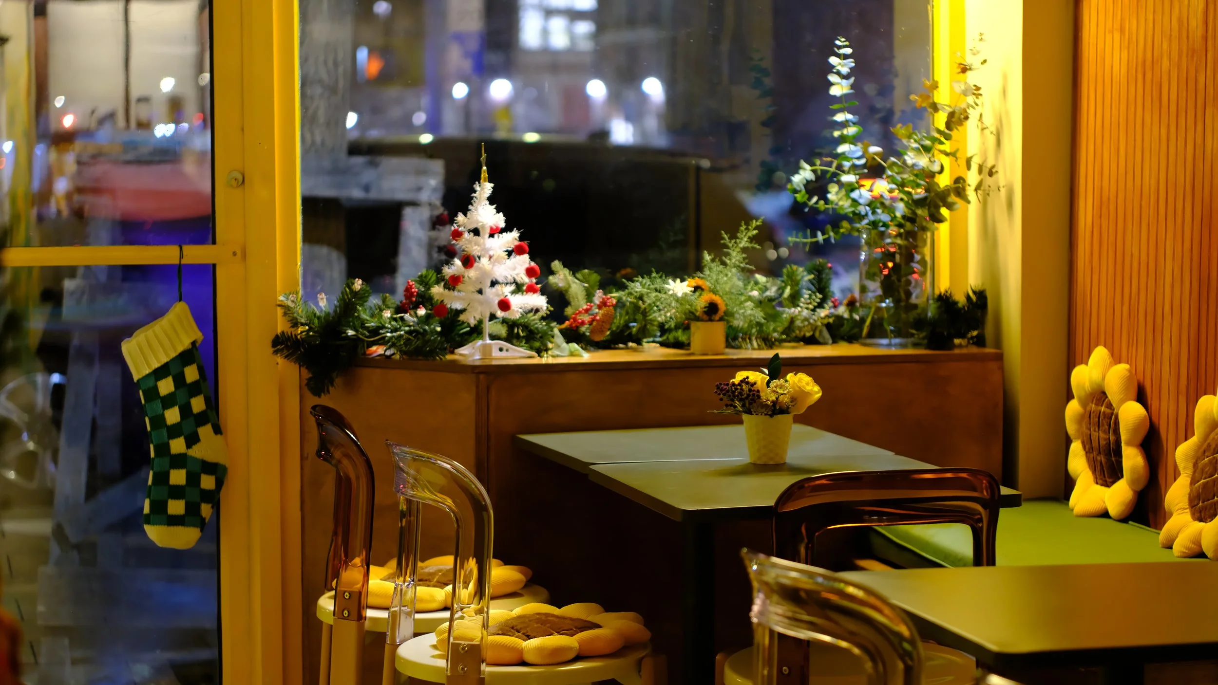 Cozy cafe decorated for Christmas with small white Christmas tree with red ornaments, various green plants, and yellow cushions on chairs.